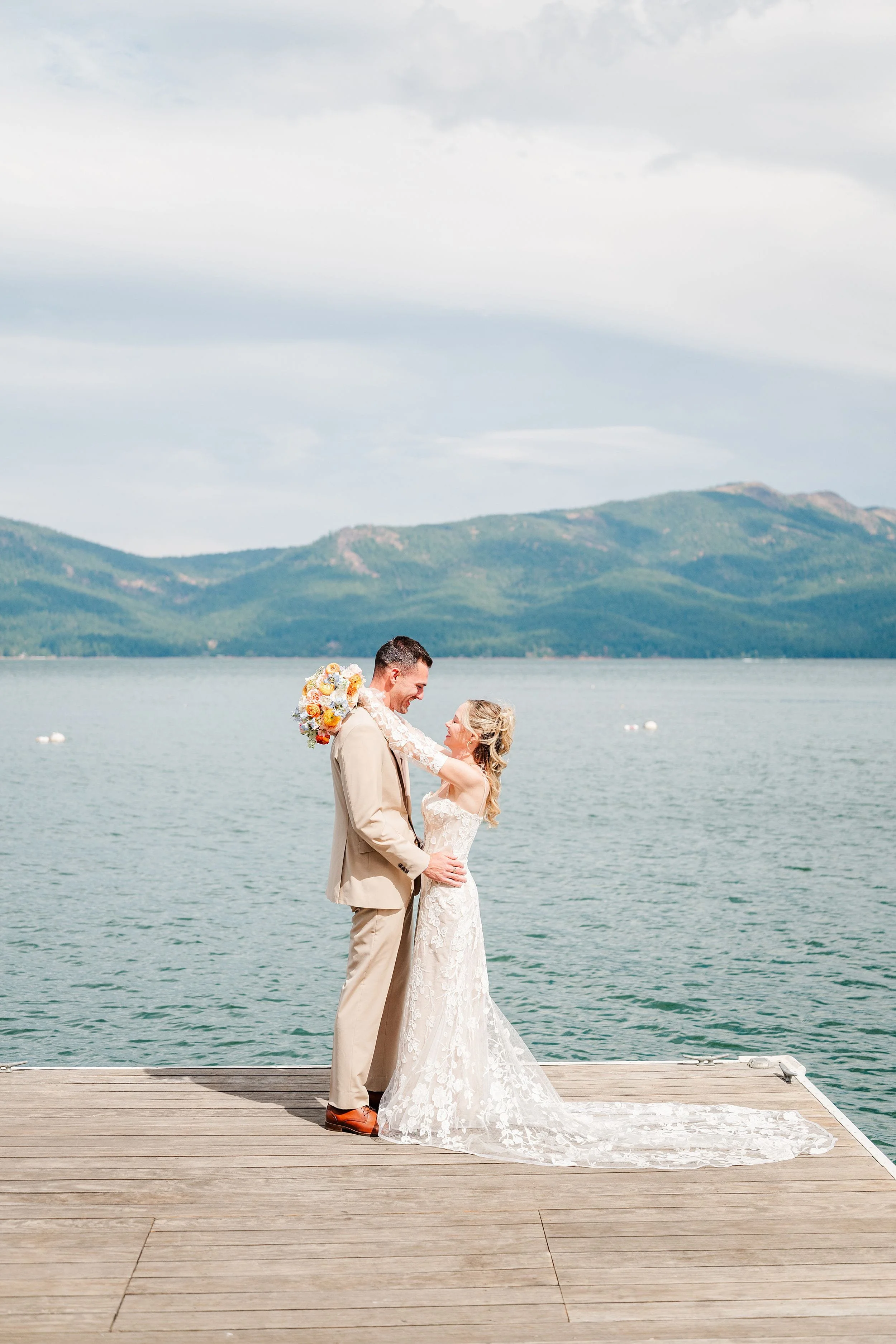 Newlywed couple embraces on a wooden dock over a mountain lake, bride in a lace gown and groom in a tan suit, creating a serene, romantic scene.