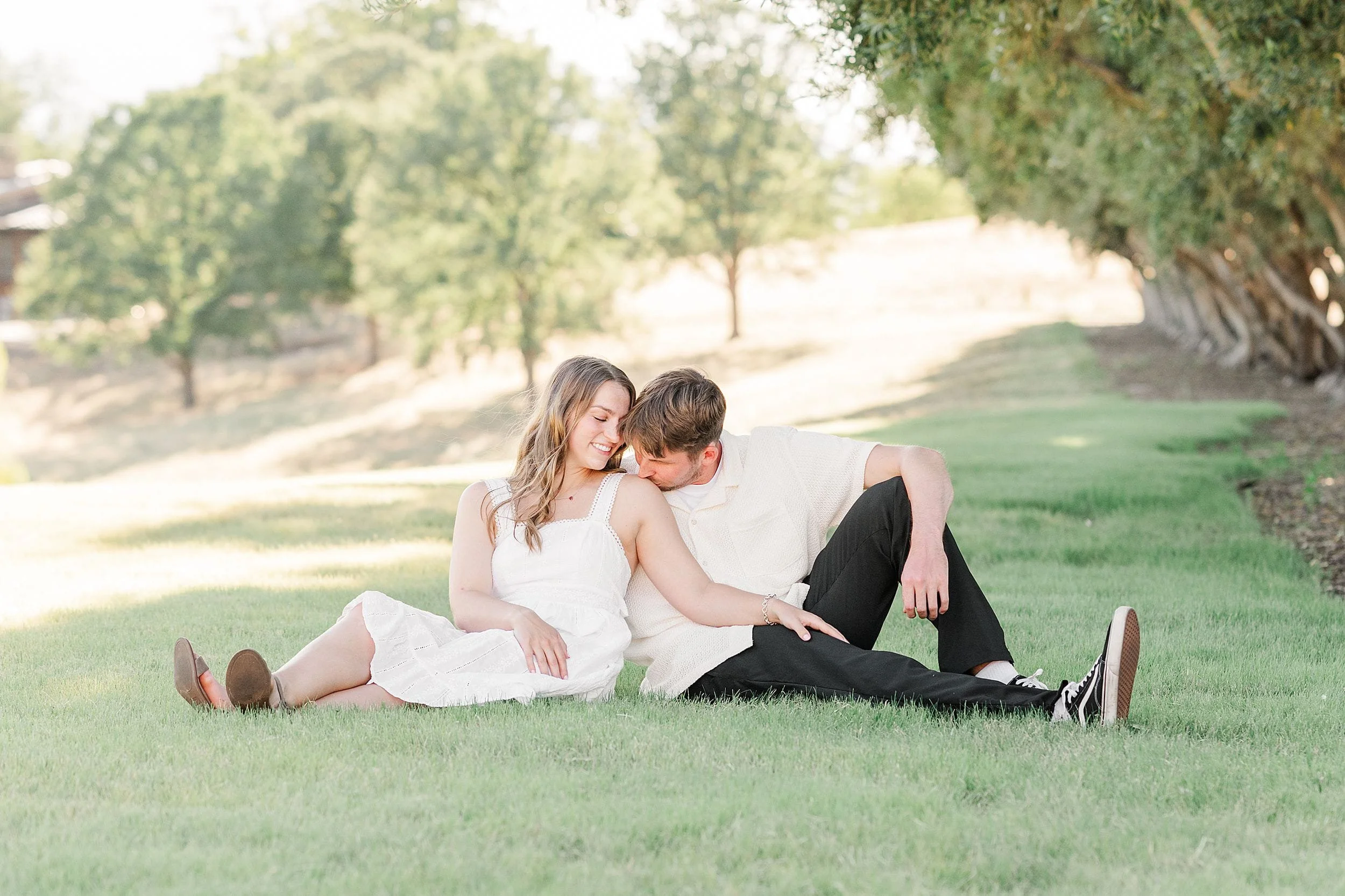 A couple sits on a sunlit grassy field, embracing affectionately. They're under trees, sharing a peaceful, intimate moment. The atmosphere is serene and romantic.