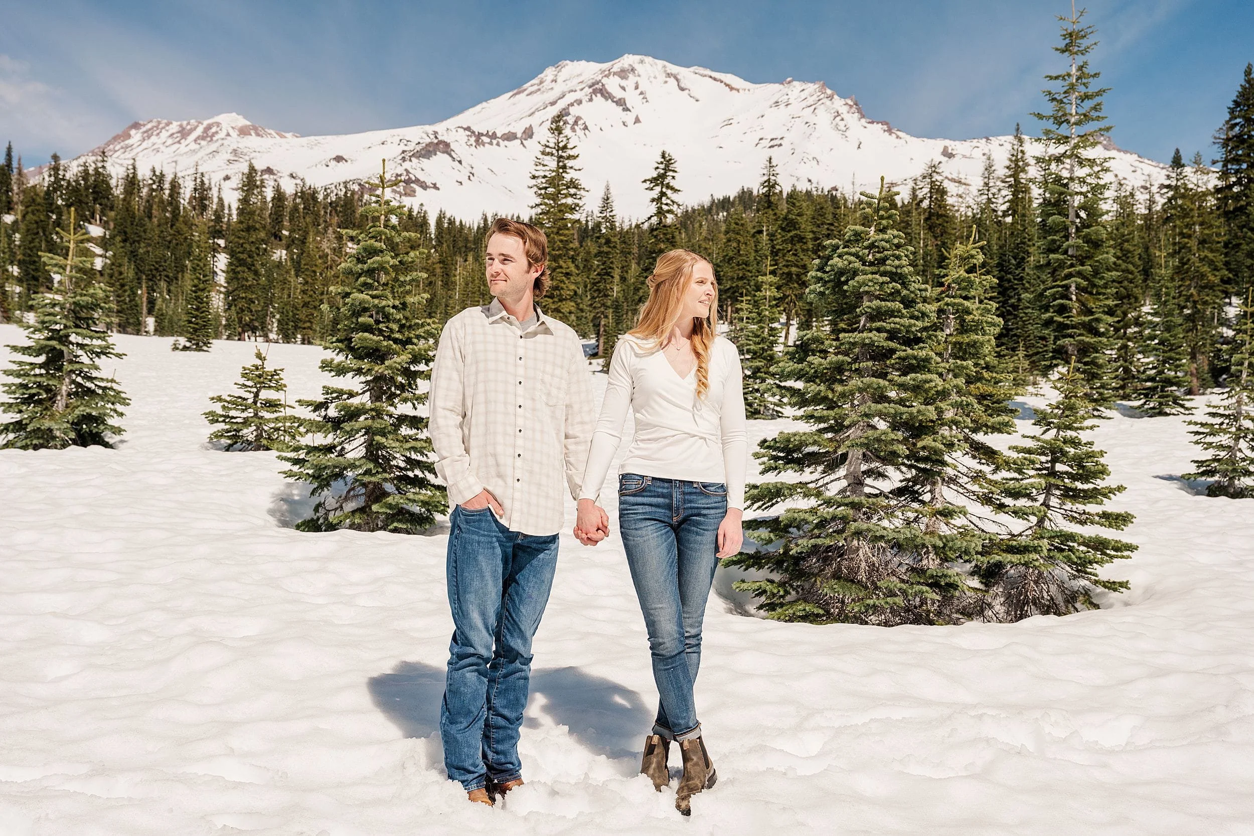 A couple in casual attire holds hands, standing in a snowy landscape with evergreen trees and a mountain backdrop under a clear blue sky.