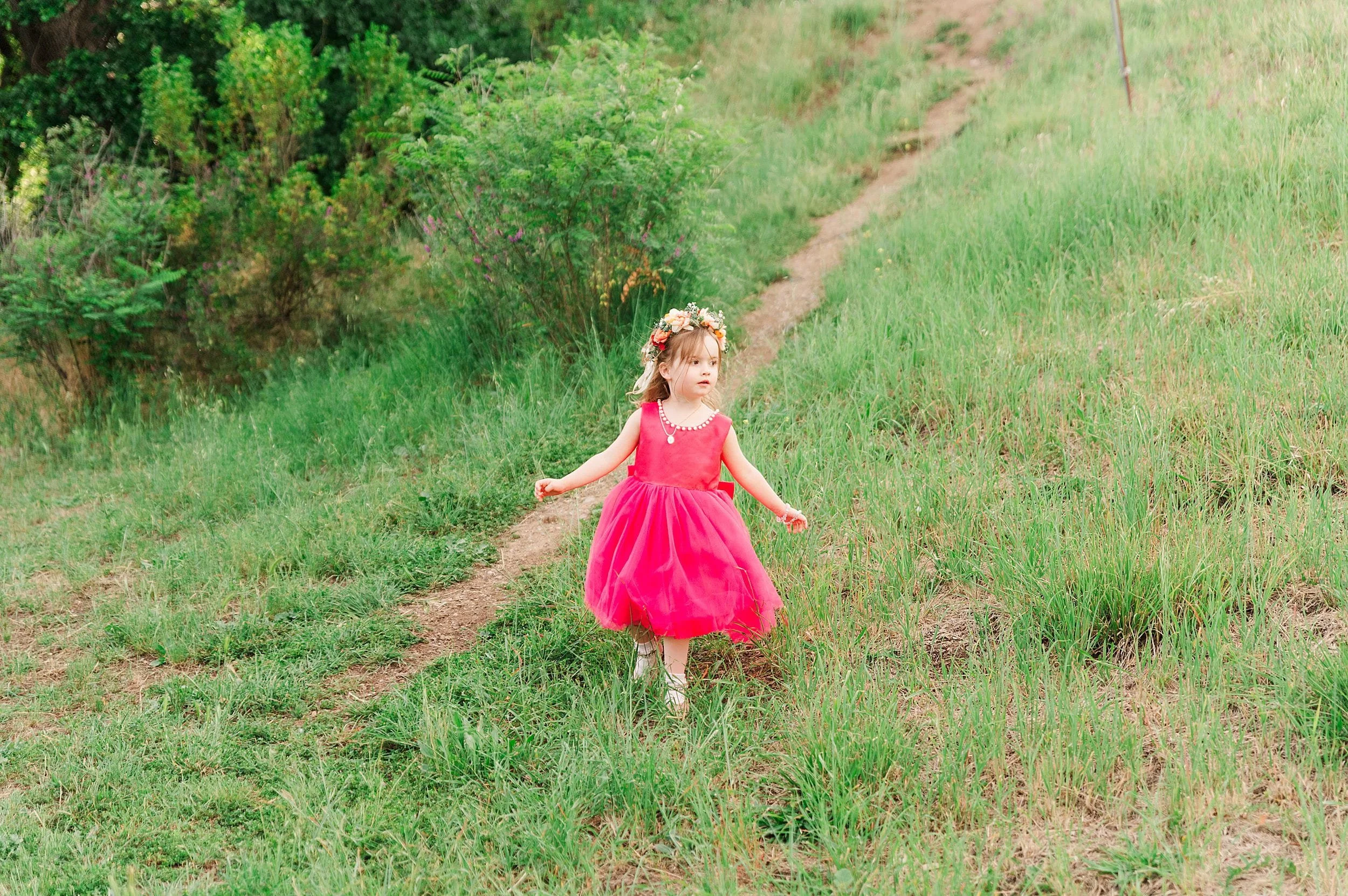 A young girl in a bright pink dress and floral crown walks along a grassy path. She explores with a curious expression amid lush green surroundings.