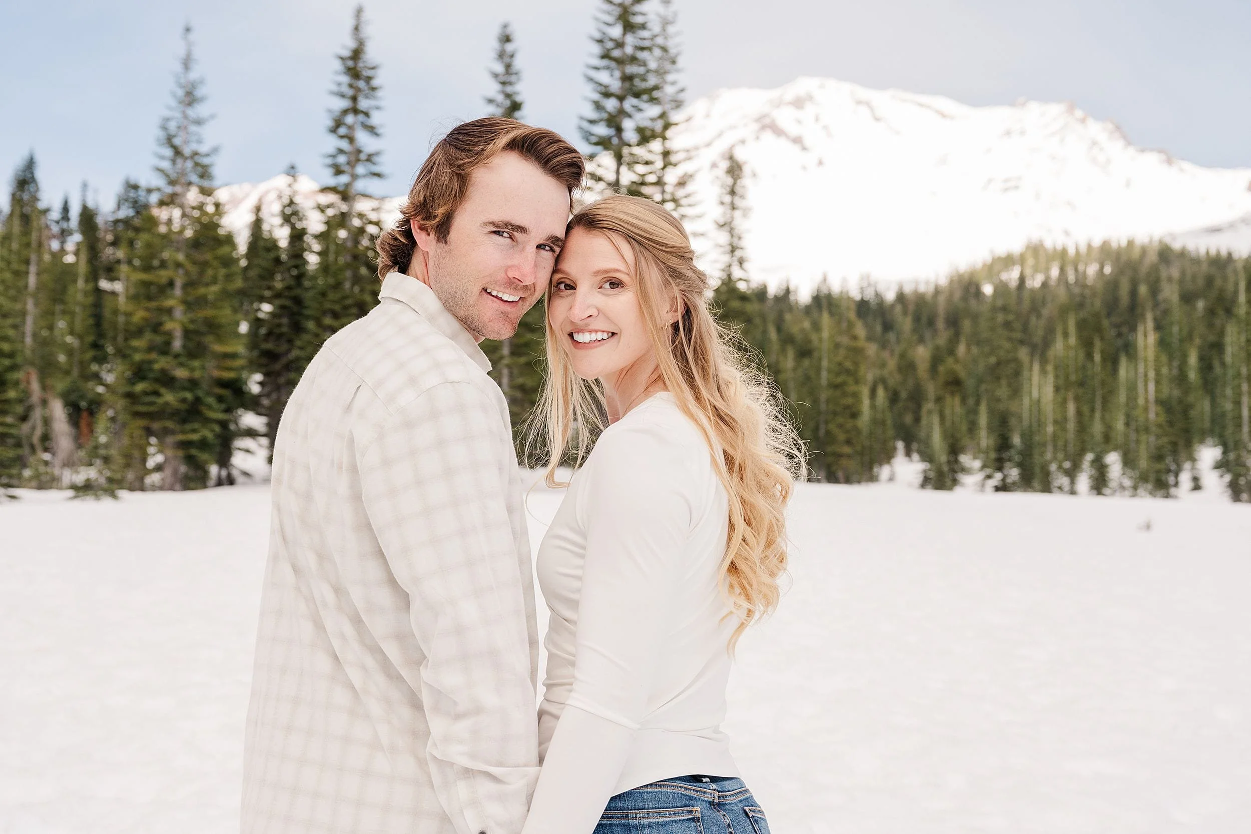 A smiling couple embraces in a snowy landscape, surrounded by pine trees and mountains. The sky is clear, conveying a serene and joyful mood.