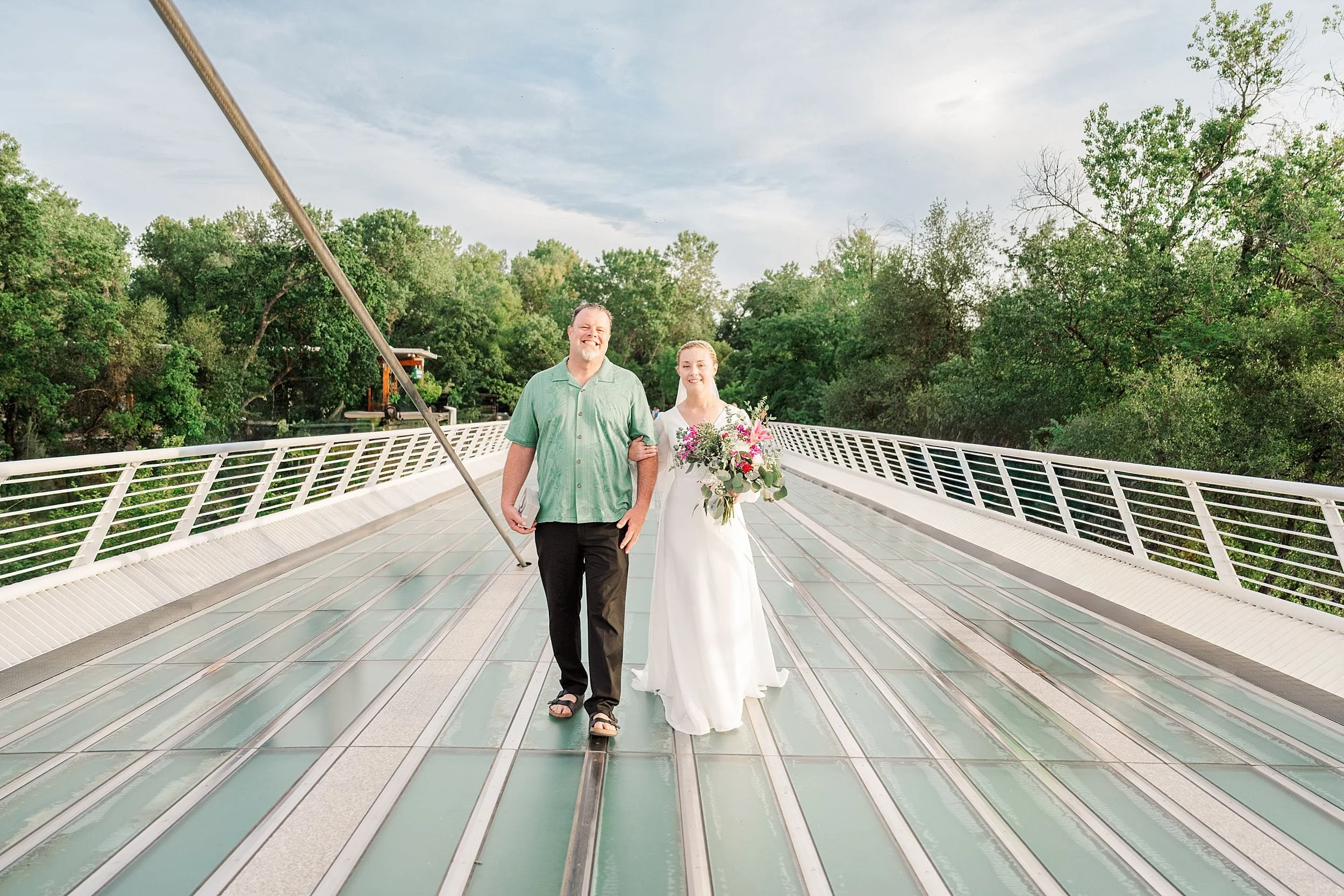 A smiling bride in a white dress holds a colorful bouquet, walking arm-in-arm with a man in a green shirt on a modern bridge, surrounded by trees.