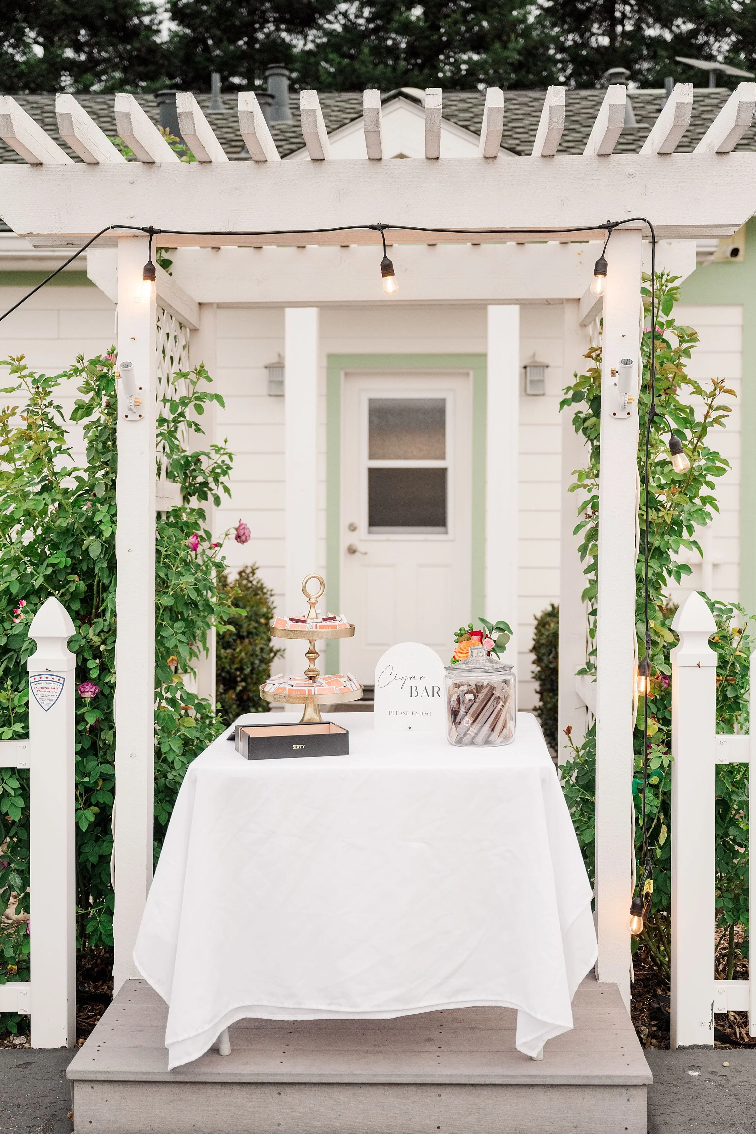 White pergola with string lights frames a small table covered in a white cloth, topped with desserts and a jar labeled "Cigar Bar." Cozy, inviting atmosphere.
