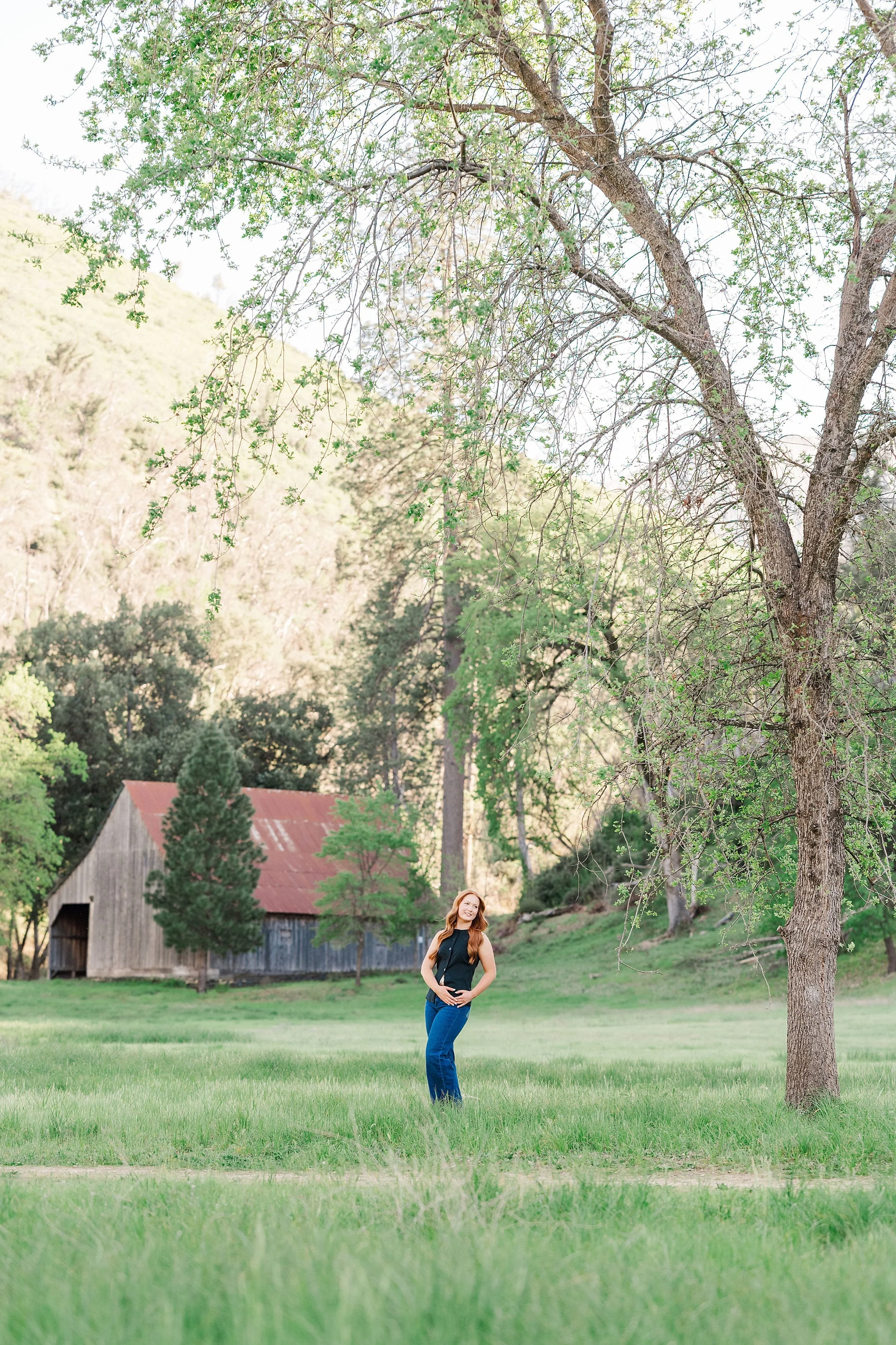 A woman stands in a lush, green field under a large tree. She wears a black top and blue jeans. In the background, there's a rustic barn with a red roof, surrounded by dense trees. The scene conveys a serene, pastoral setting.