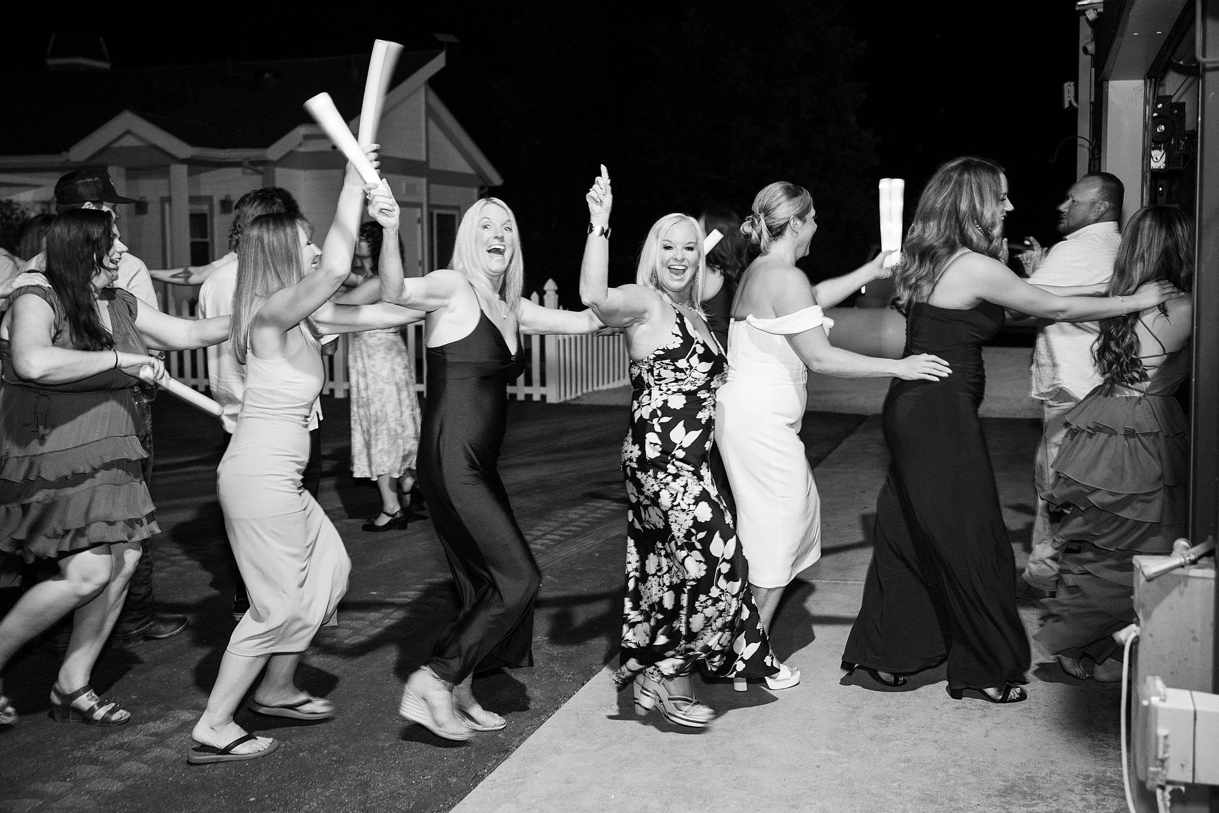 A joyful conga line of people in elegant attire dances outside at night, holding light sticks. Smiling, they exude a lively and festive atmosphere.