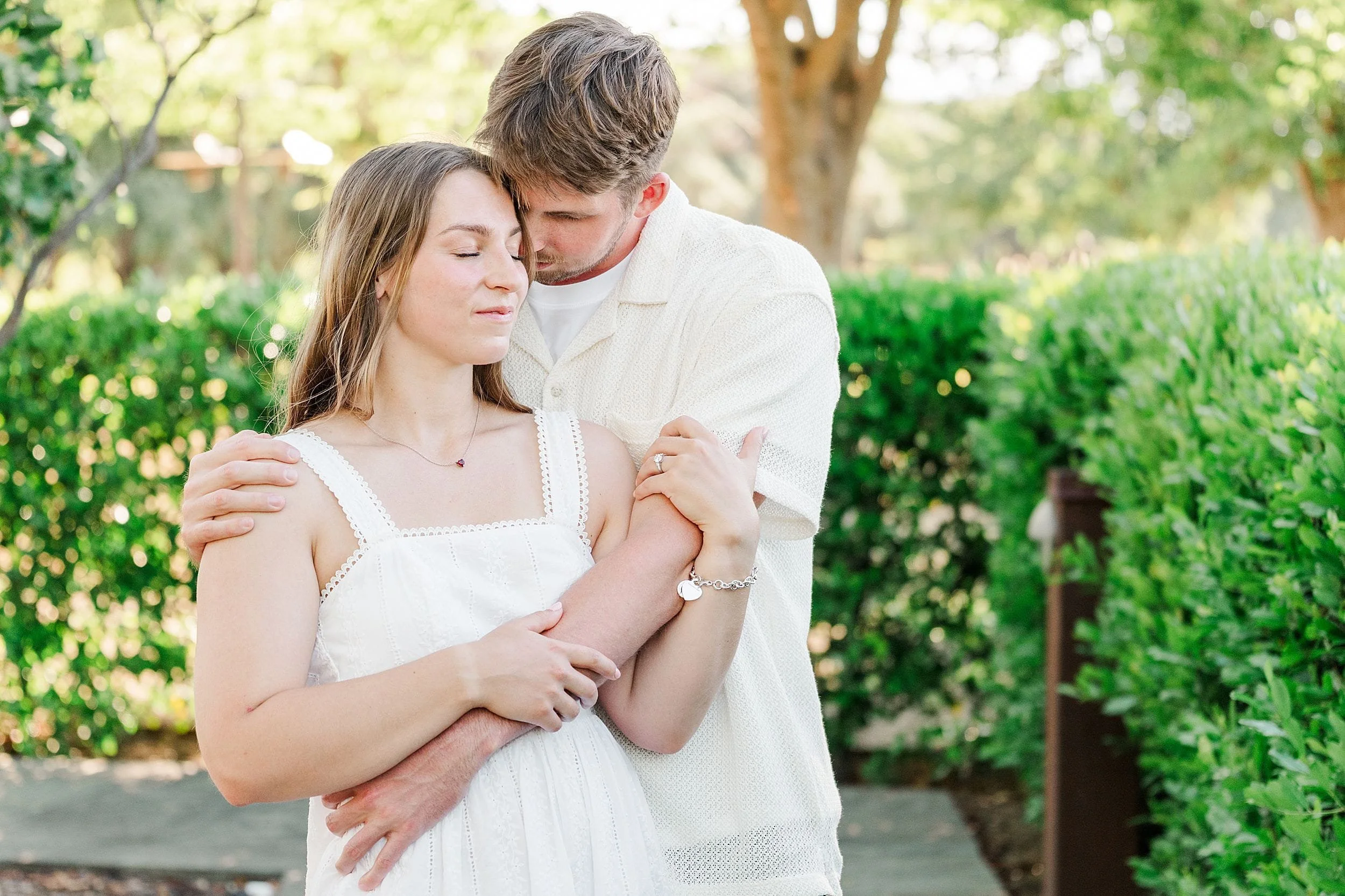 A couple embraces in a lush garden, the woman in a white dress with closed eyes and a serene expression. The man, in a white shirt, gently hugs her, conveying tenderness.