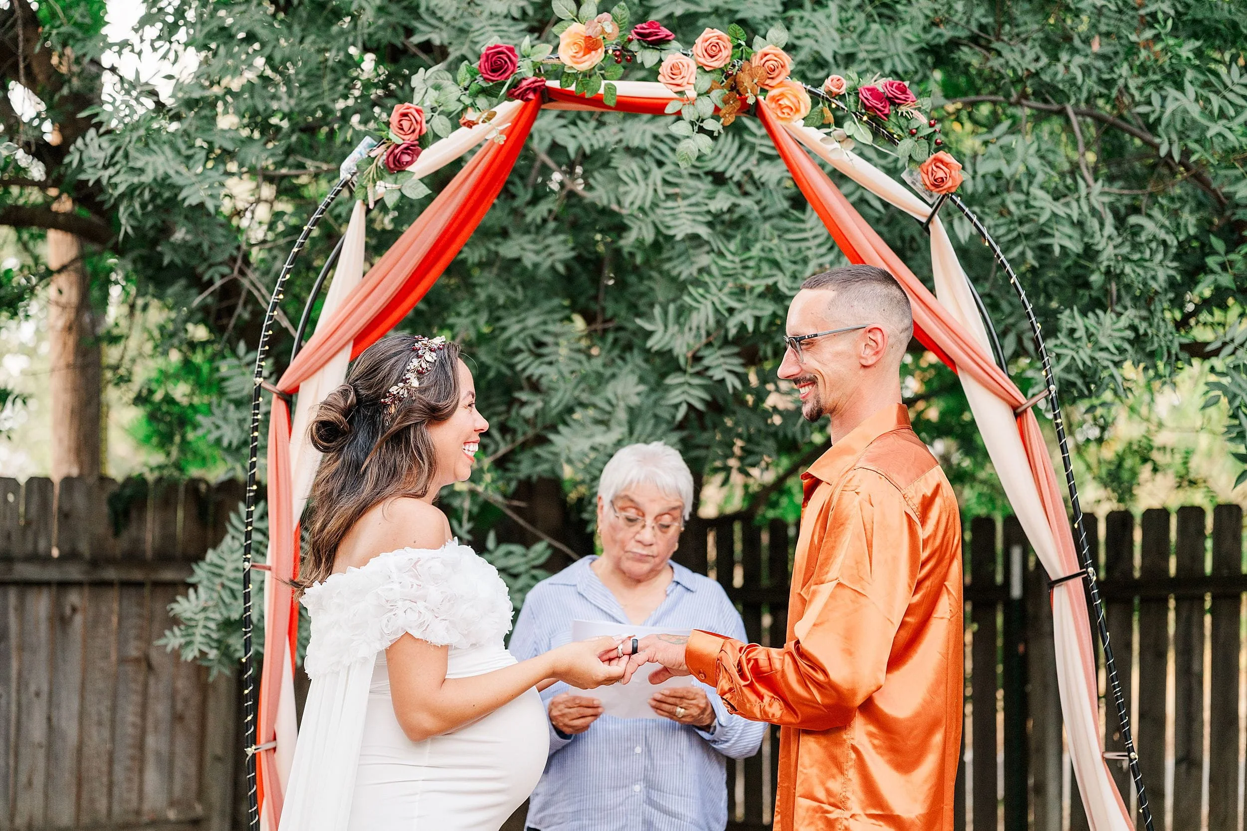 Bride and groom exchange rings beneath a floral arch draped in red and ivory fabric, while an officiant oversees the joyful outdoor ceremony.