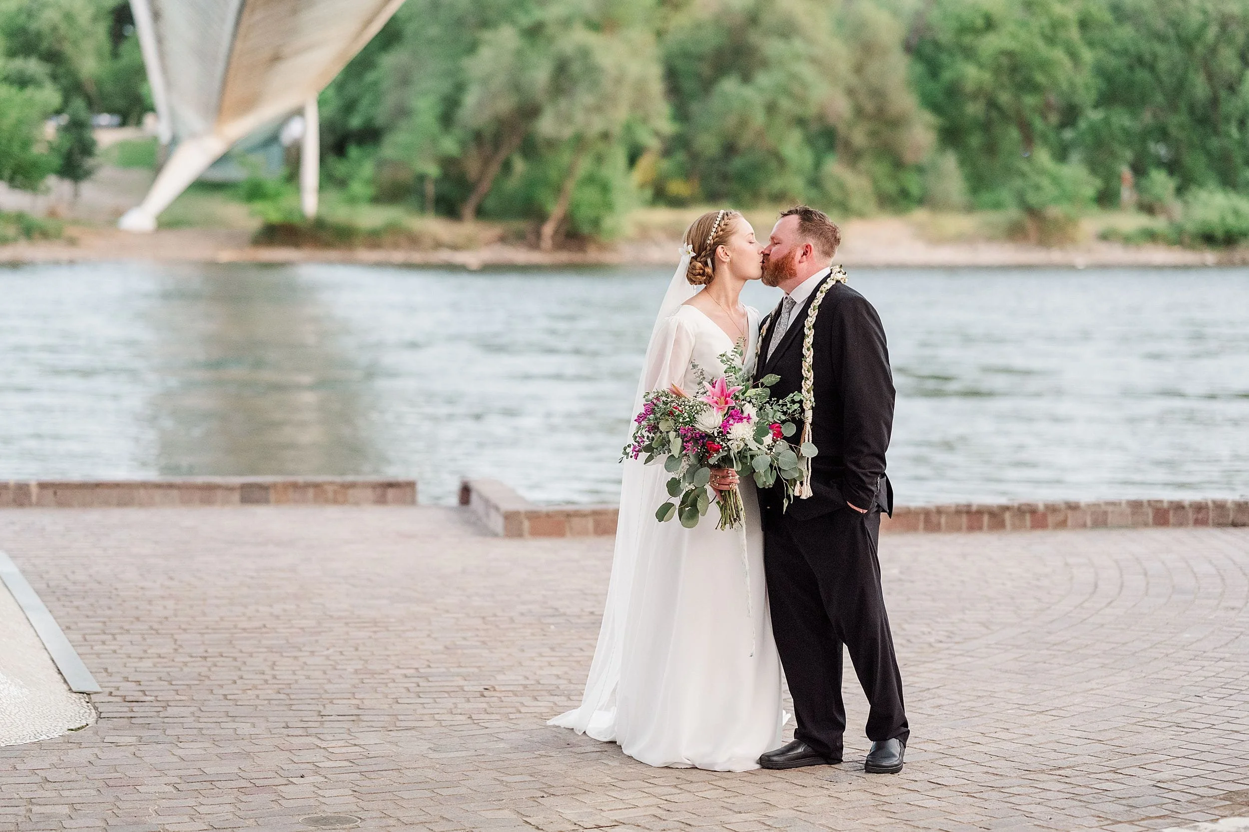 A bride and groom share a kiss by a river, under a modern bridge. The bride holds a vibrant bouquet, and greenery surrounds the scene, conveying romance.