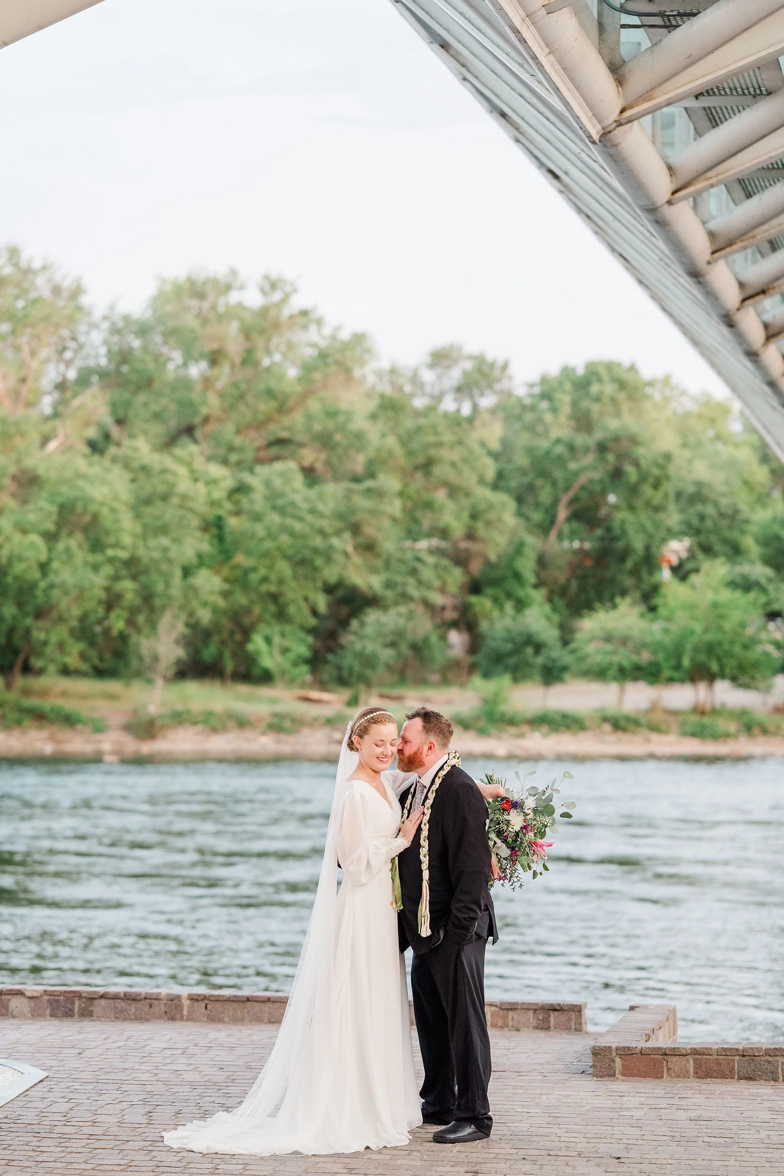 A couple in wedding attire stands by a river, framed by lush greenery and a modern architectural structure. The scene conveys joy and serenity.