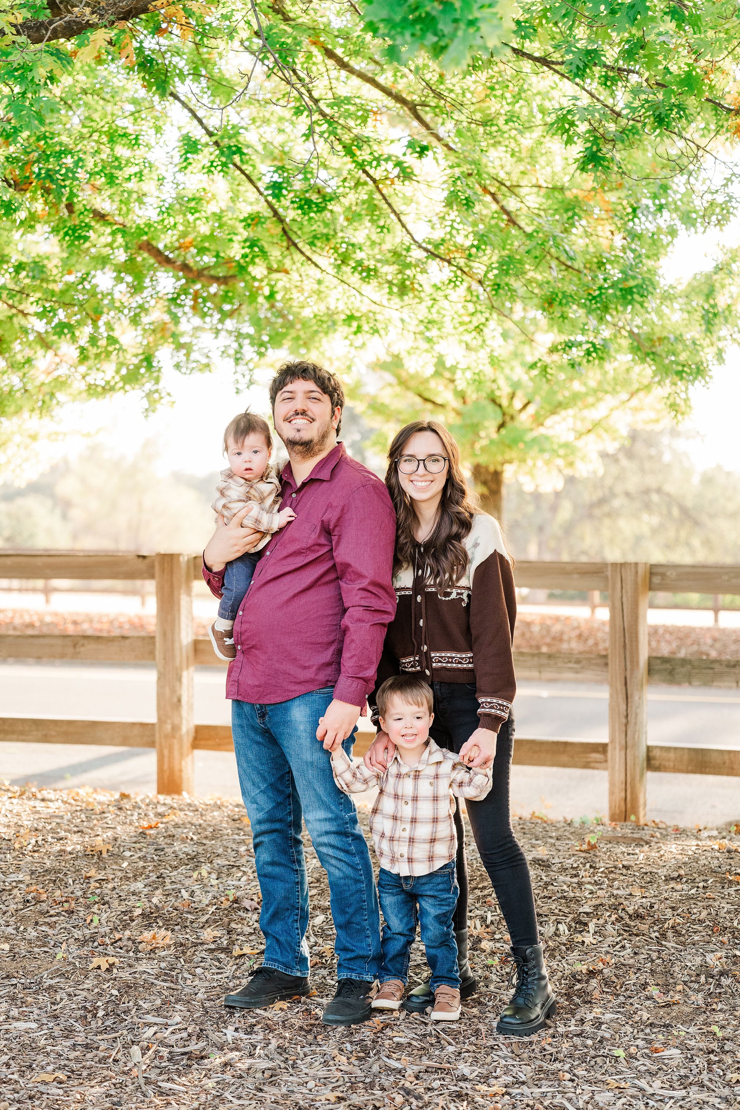 A smiling family of four poses beneath a leafy tree beside a wooden fence, creating a warm, relaxed portrait in dappled sunlight.
