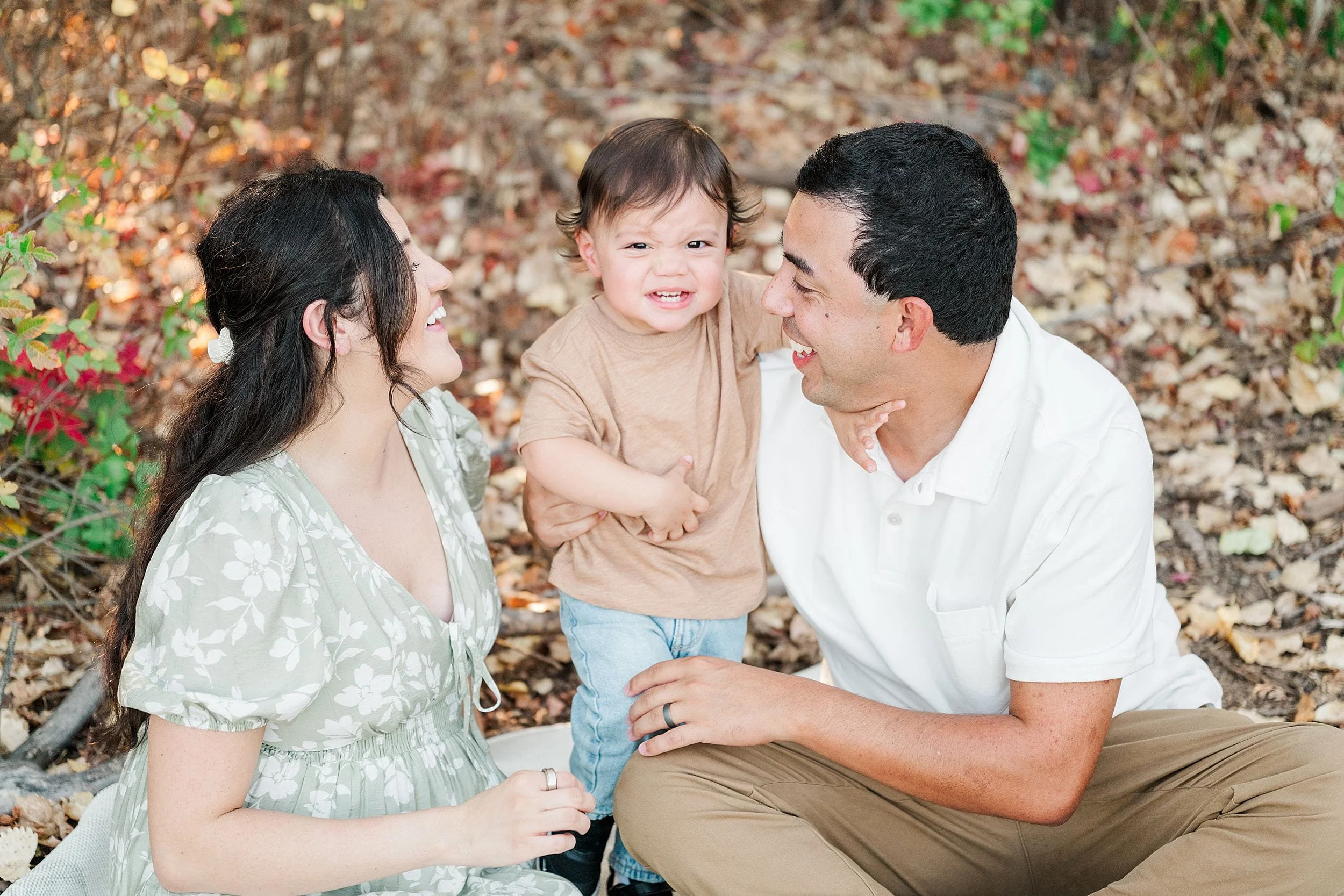 A smiling toddler stands between his parents in a leafy autumn setting, as they gaze at him with affection; the family portrait feels warm and joyful.