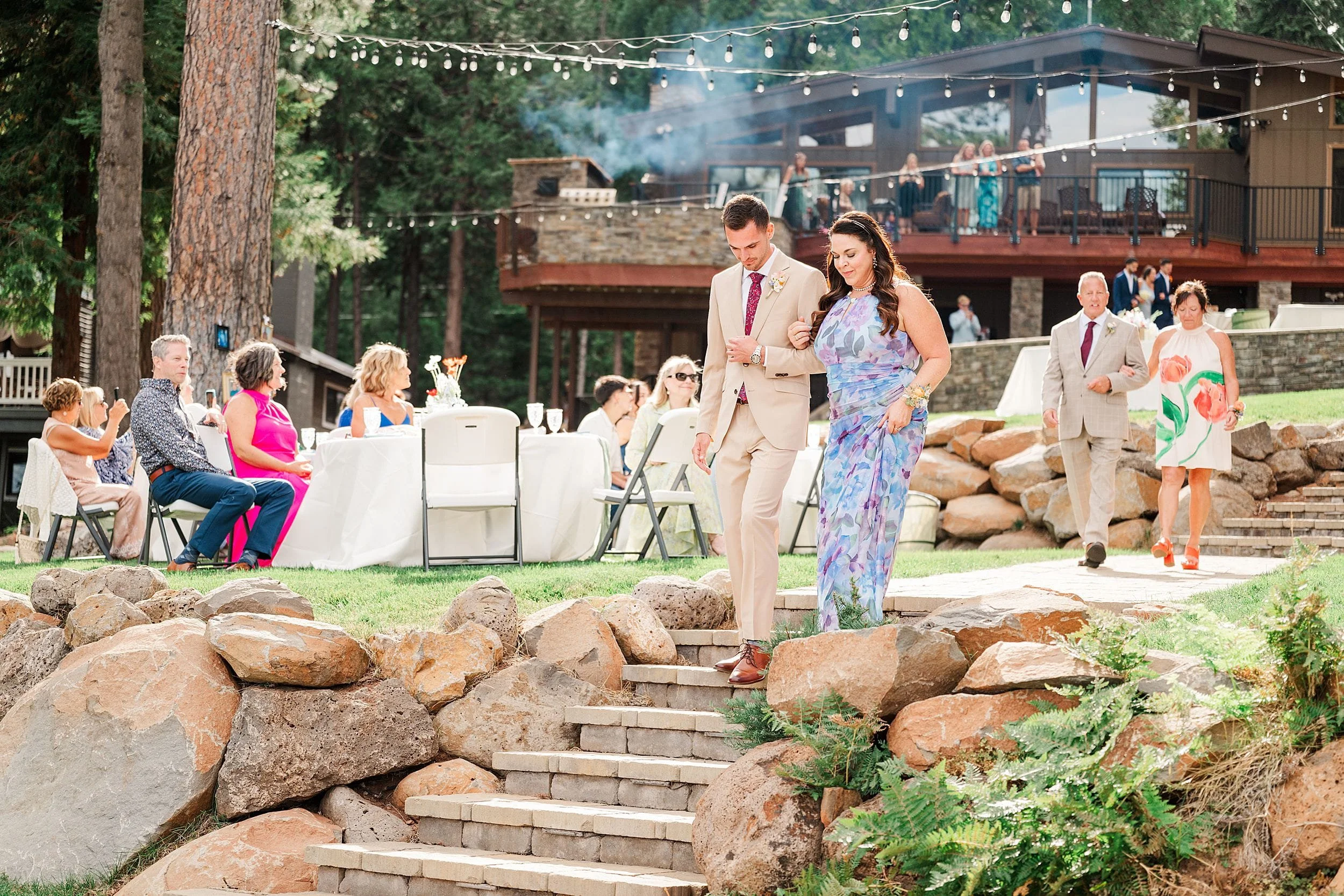 Groom and his mom descends stone steps at a rustic outdoor wedding venue, surrounded by smiling guests, string lights, trees, and warm sunlight.