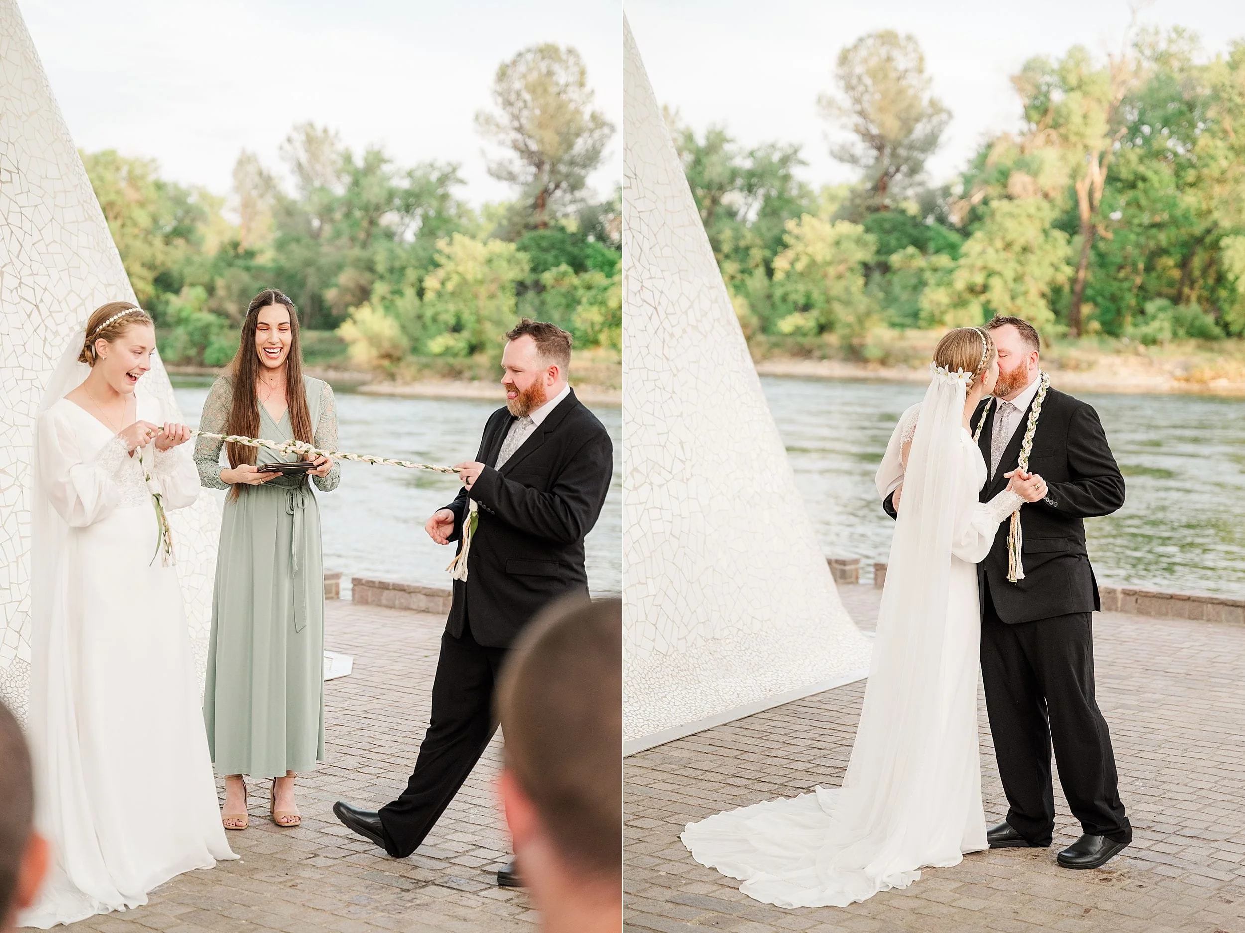 A couple in wedding attire joyfully participates in a handfasting ceremony by a river. They are smiling, surrounded by greenery and a white, triangular backdrop.