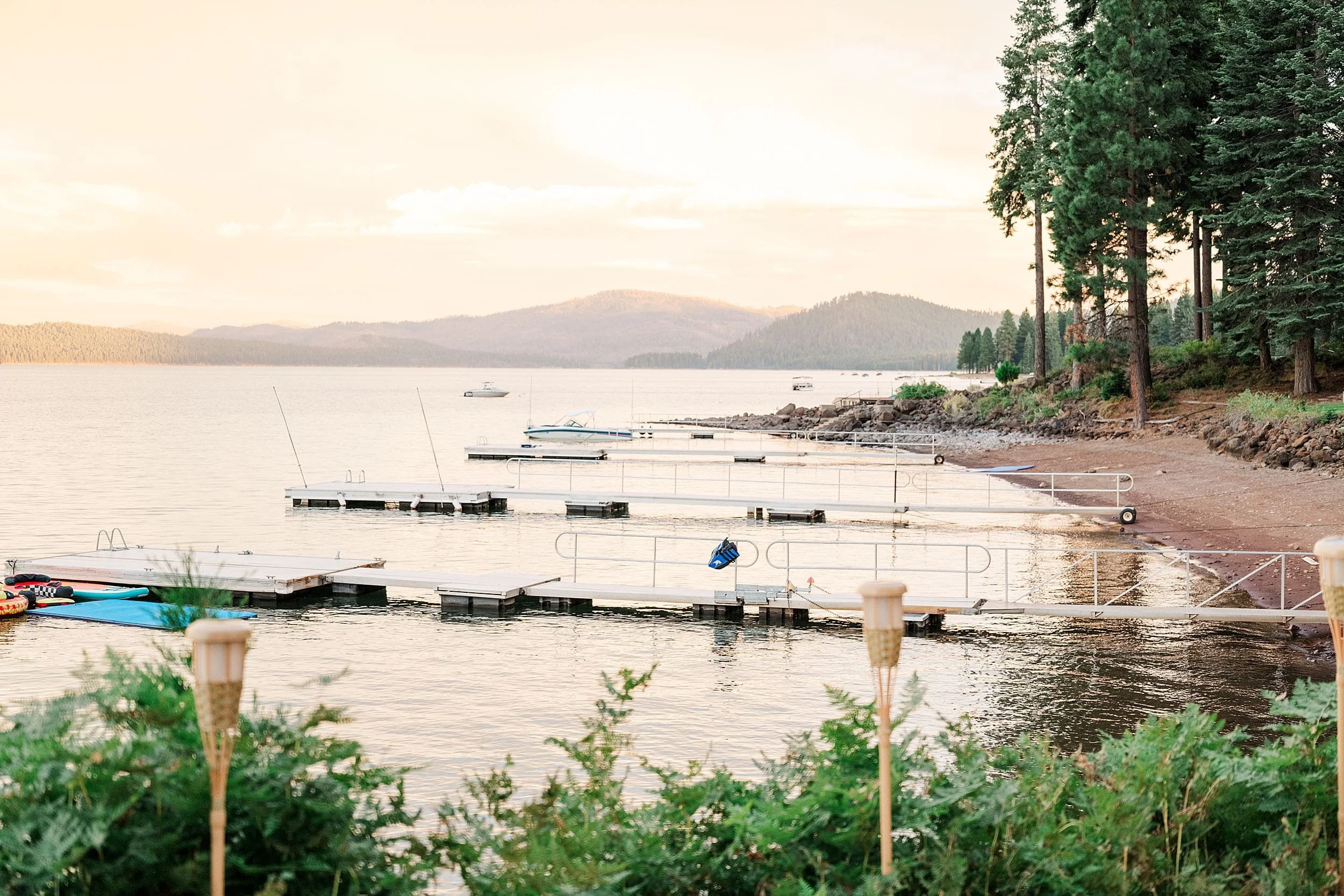 Quiet lakeside marina with empty floating docks, kayaks, forested shoreline, and hazy mountains under a warm, peaceful sky.