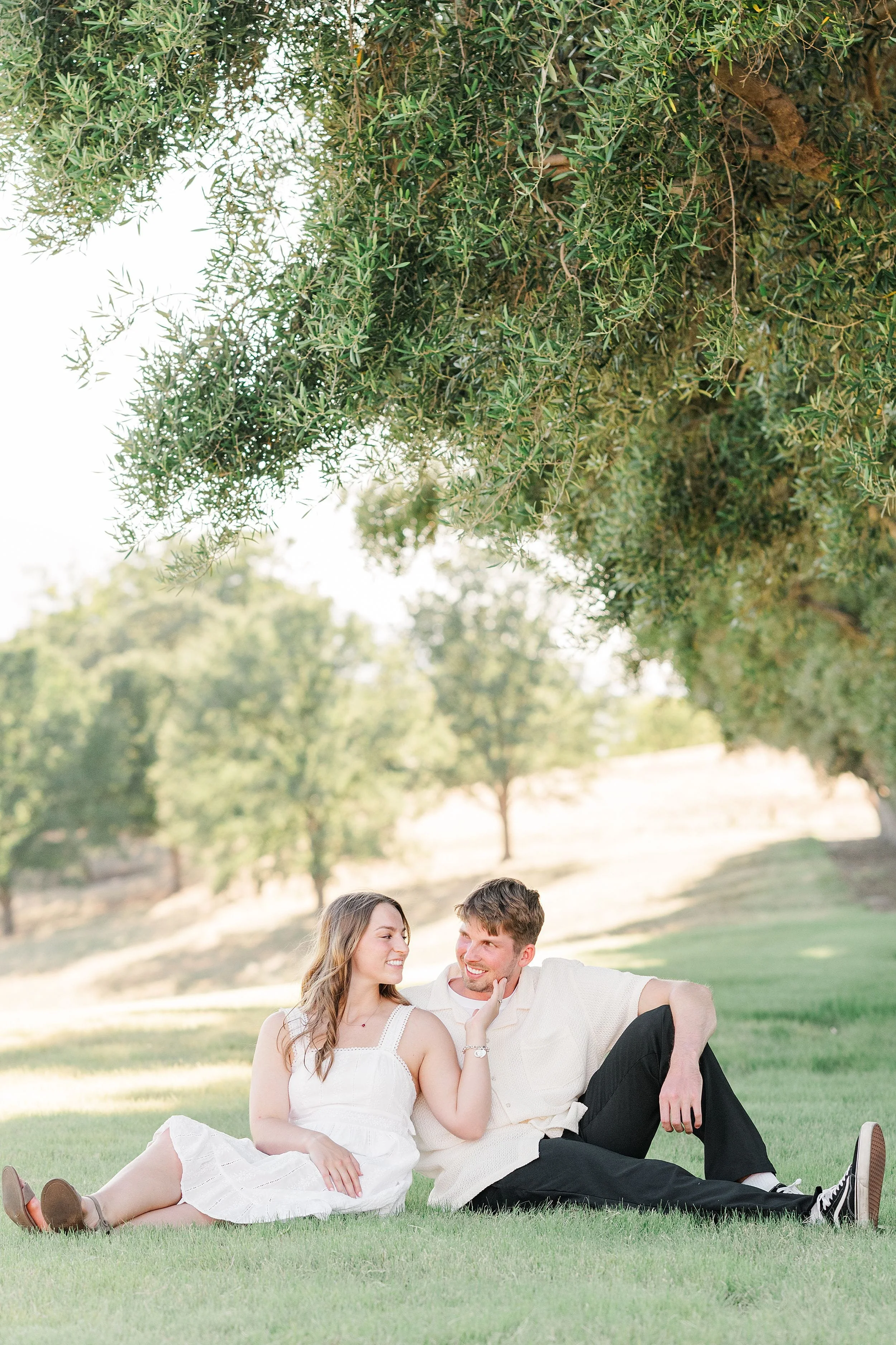 A couple in casual summer outfits sits on lush grass under a large tree, sharing a joyful moment. Sunlight filters through leaves, creating a serene, romantic atmosphere.