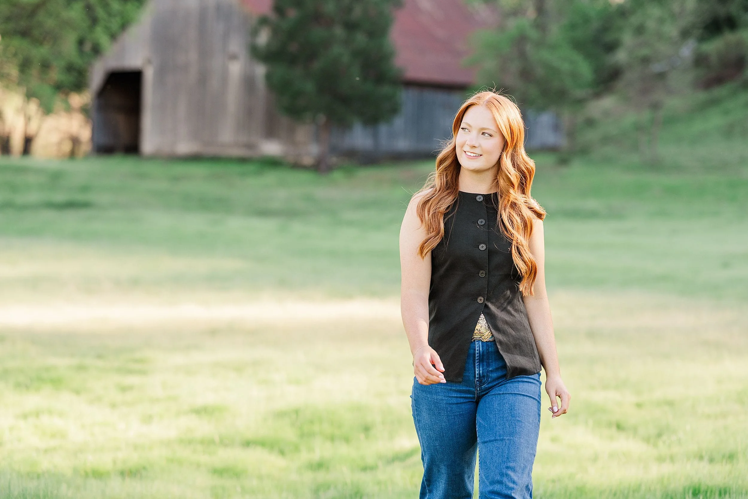 A woman with long red hair walks through a sunlit grassy field, wearing a black sleeveless top and jeans. A rustic barn stands in the background.