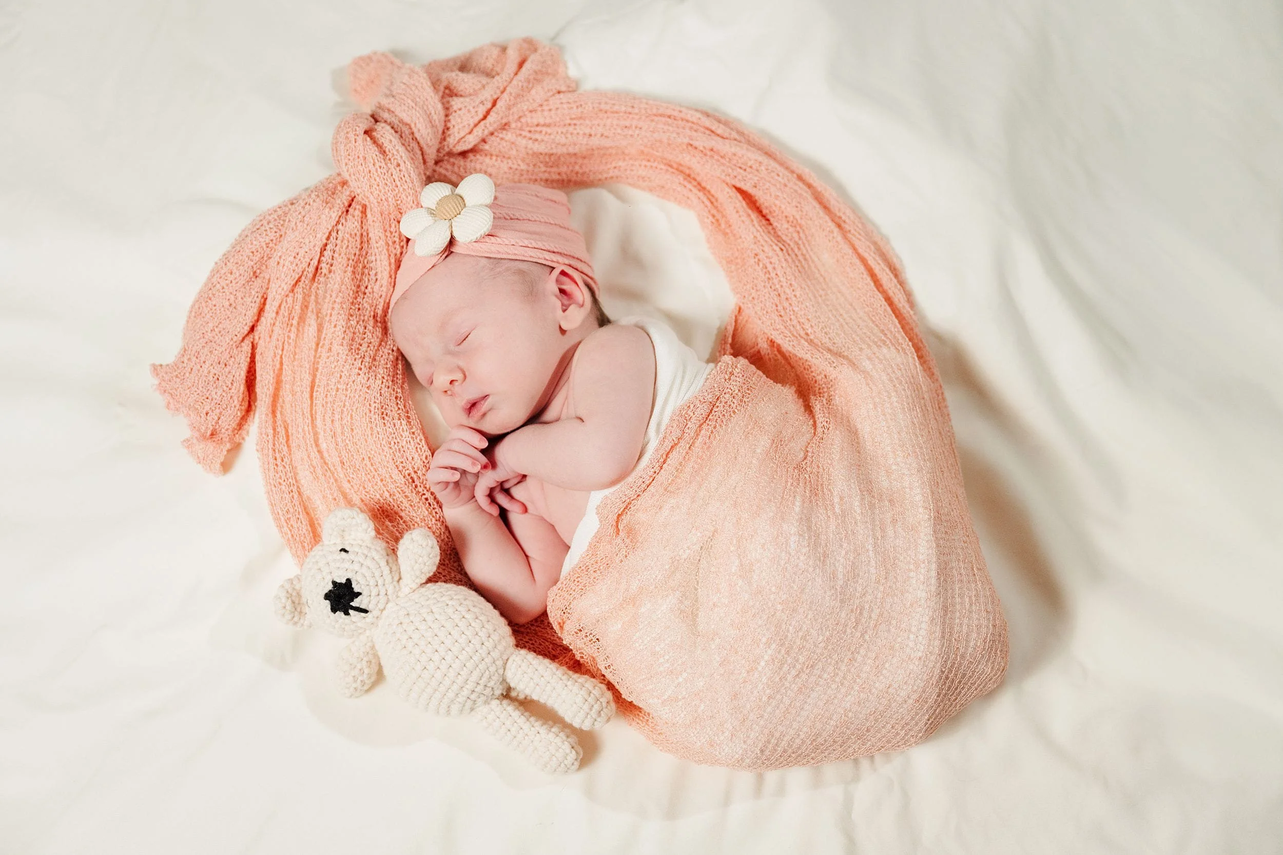 Newborn baby sleeps peacefully, wrapped in a soft, peach blanket with a matching headband adorned with flowers, next to a knitted teddy bear.