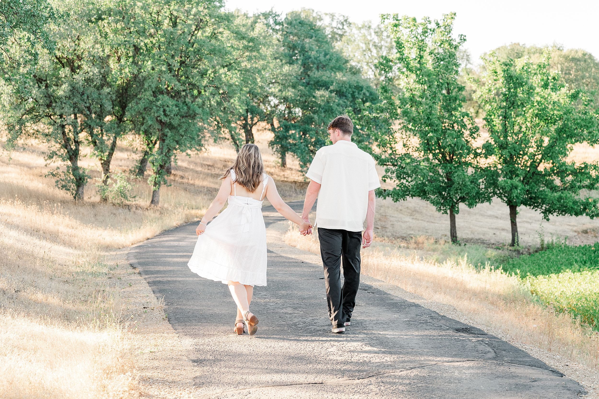 A couple walks hand in hand down a sunlit path in a rural landscape, surrounded by green trees and dry grass. The mood is serene and romantic.