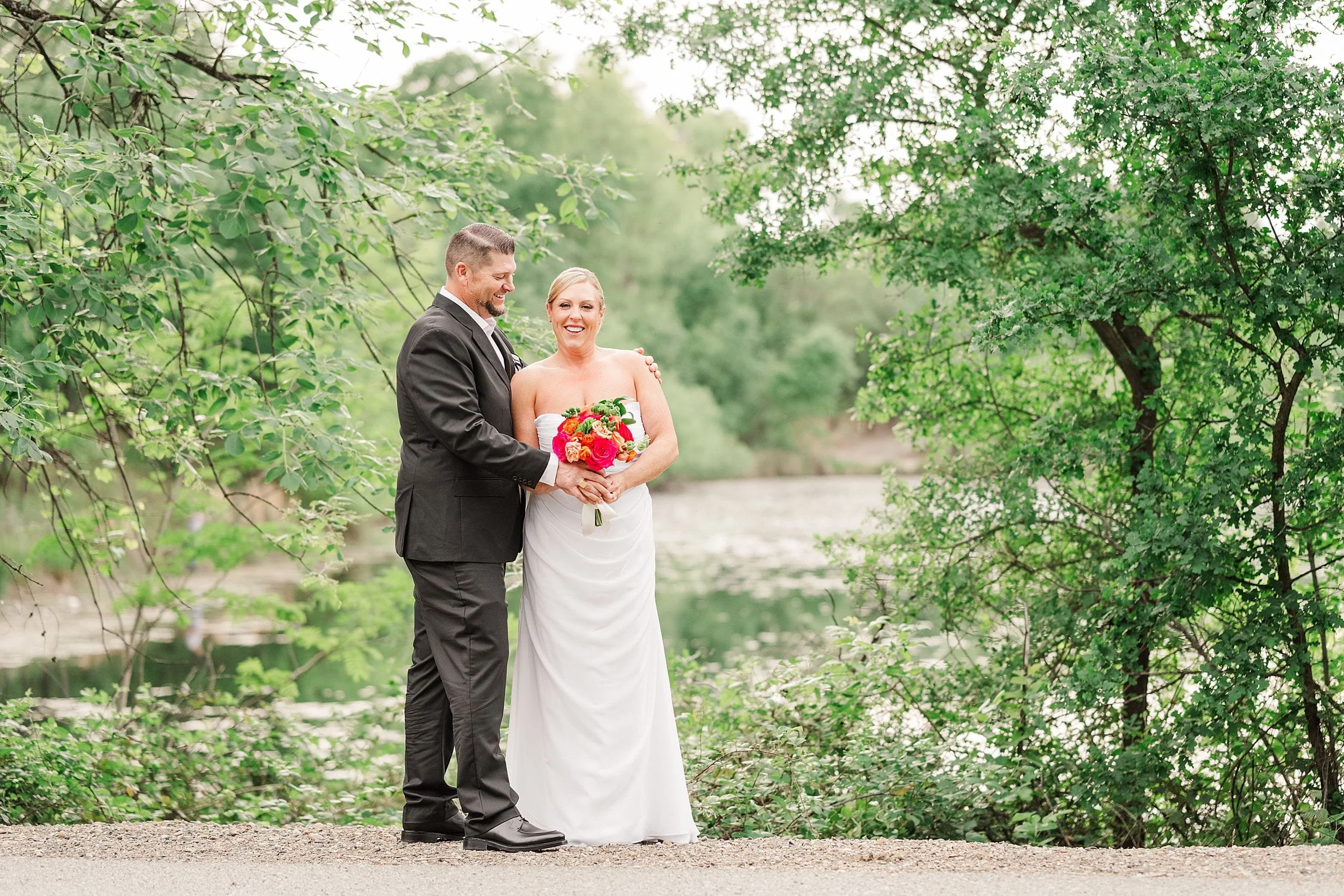 A bride in a white gown and groom in a black suit stand smiling beside a lush green riverbank. The bride holds a vibrant bouquet of flowers.