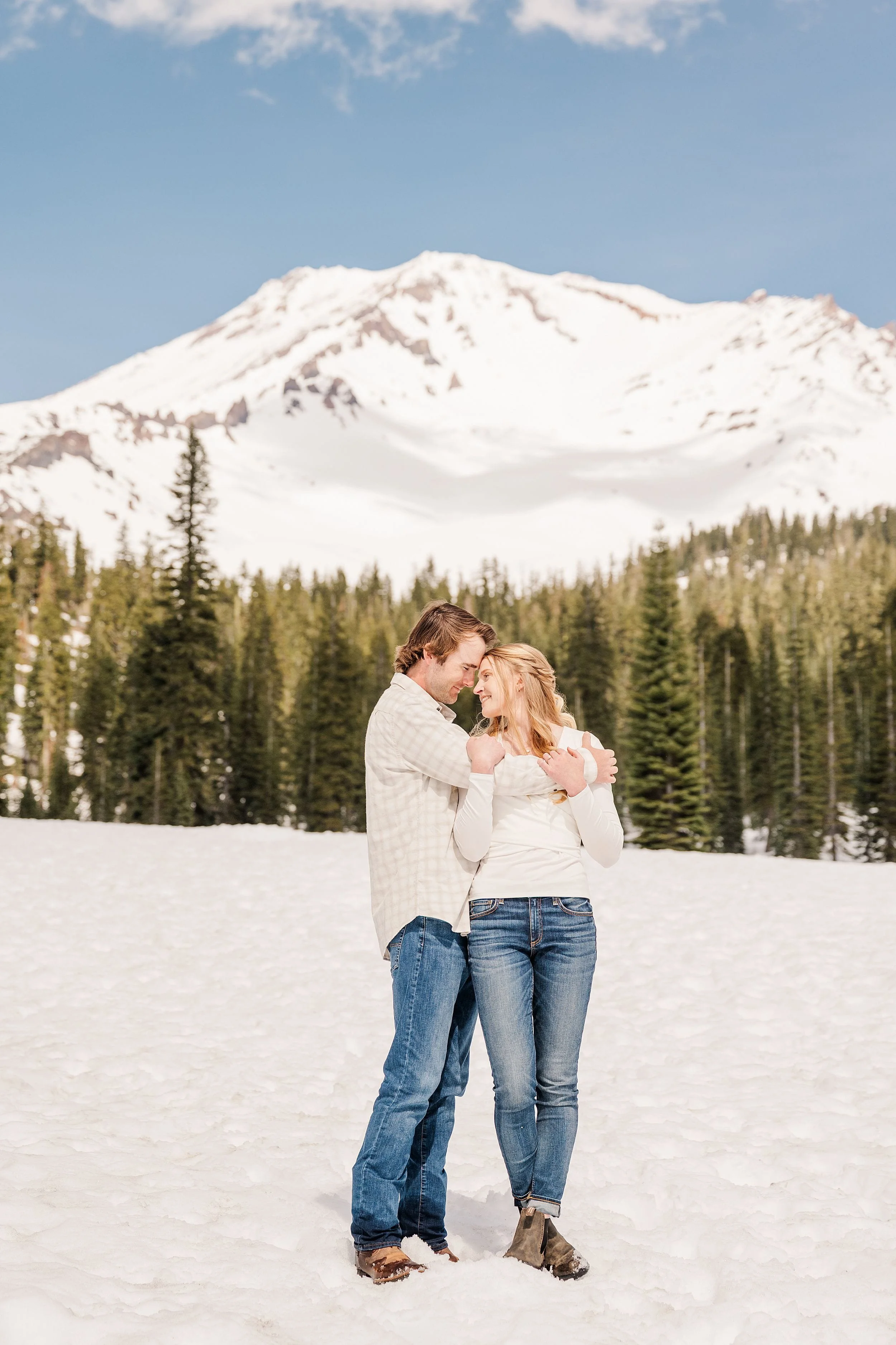 A couple stands on snow, embracing and smiling, with a backdrop of snowy mountains and evergreen trees under a clear blue sky. Romantic and joyful atmosphere.