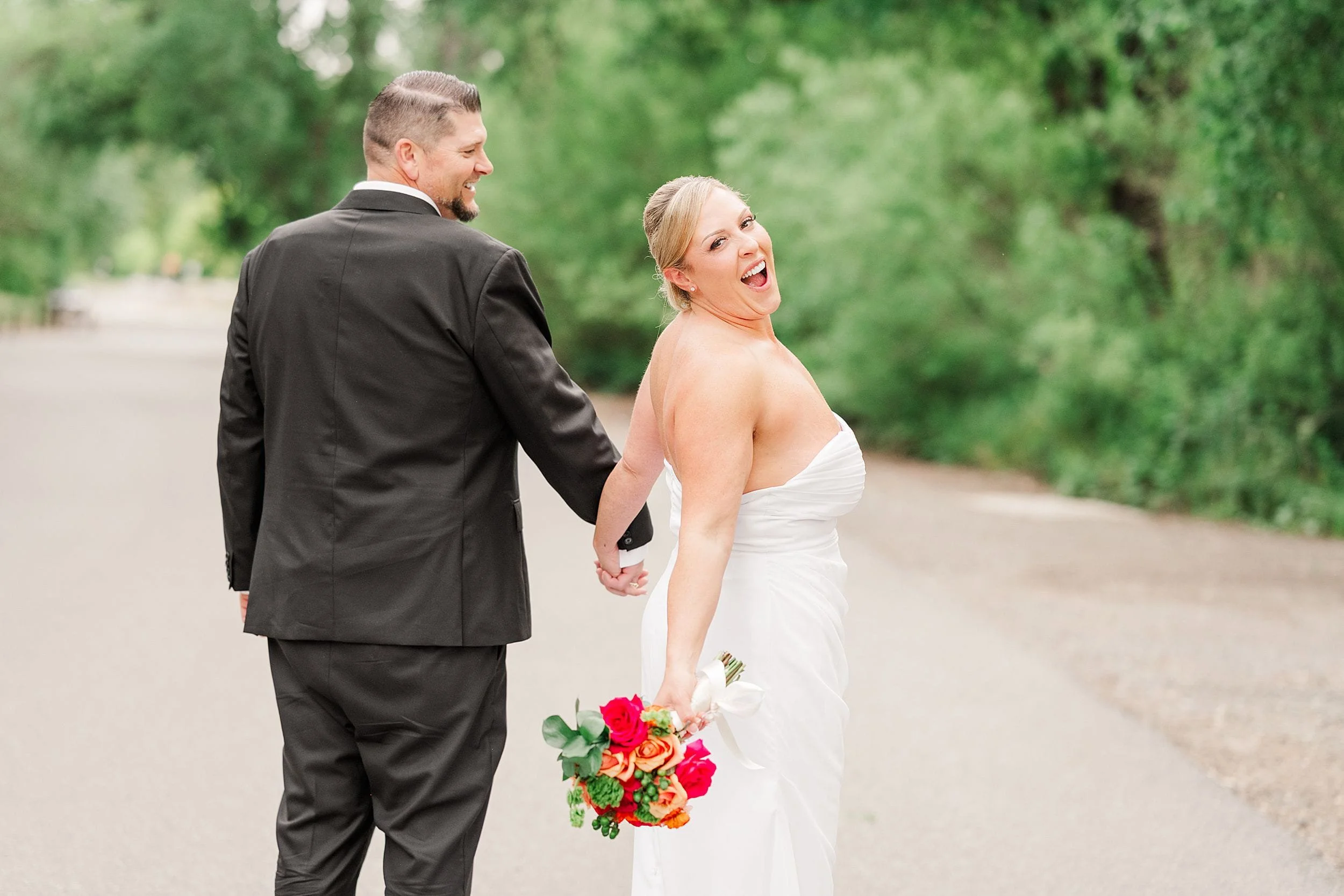 Bride in a white gown holds a colorful bouquet and the hand of a groom in a black suit. Both smile and walk along a tree-lined path, exuding joy.
