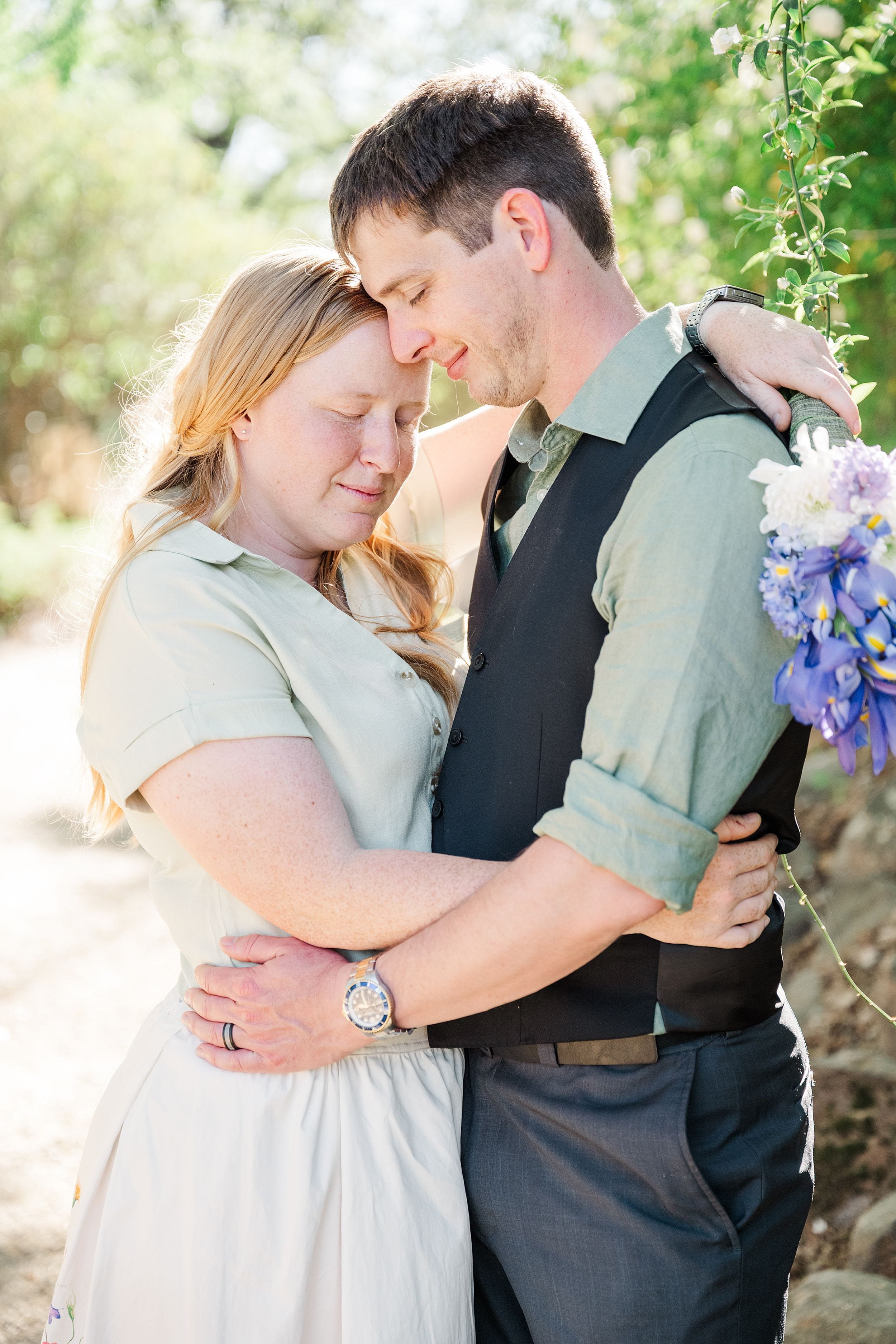 A couple embraces warmly in a sunlit garden, eyes closed and smiling softly. The woman holds a bouquet of purple flowers. The scene conveys love and serenity.