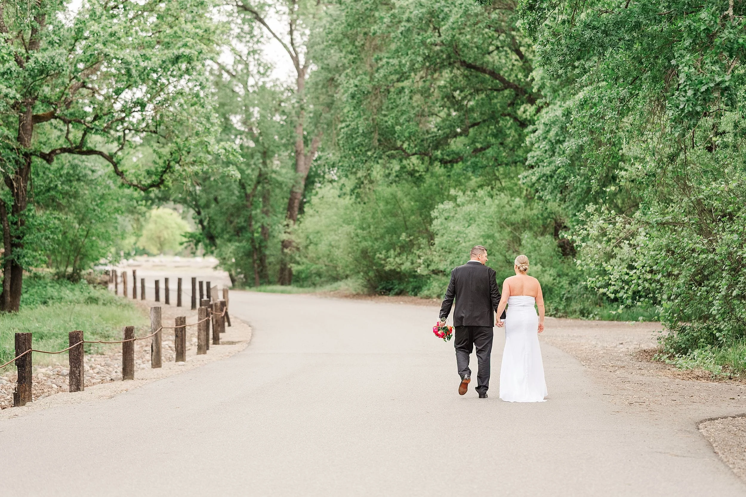 A bride and groom walk hand in hand down a tree-lined path, surrounded by lush greenery. The groom holds a bouquet, conveying a serene, romantic mood.