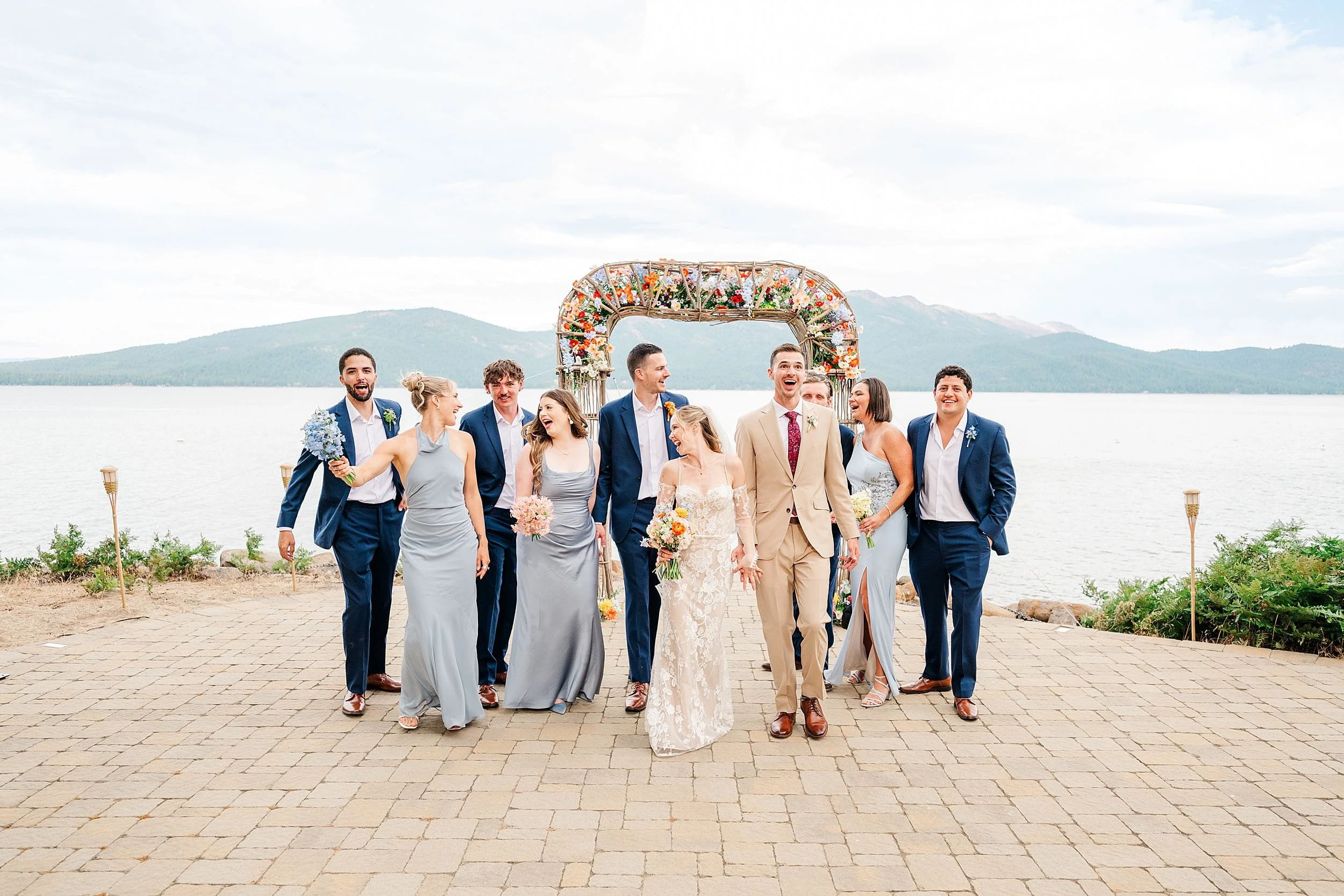 Joyful wedding party poses beneath a floral arch by a lakeside, with bride and groom surrounded by bridesmaids and groomsmen, mountains behind.