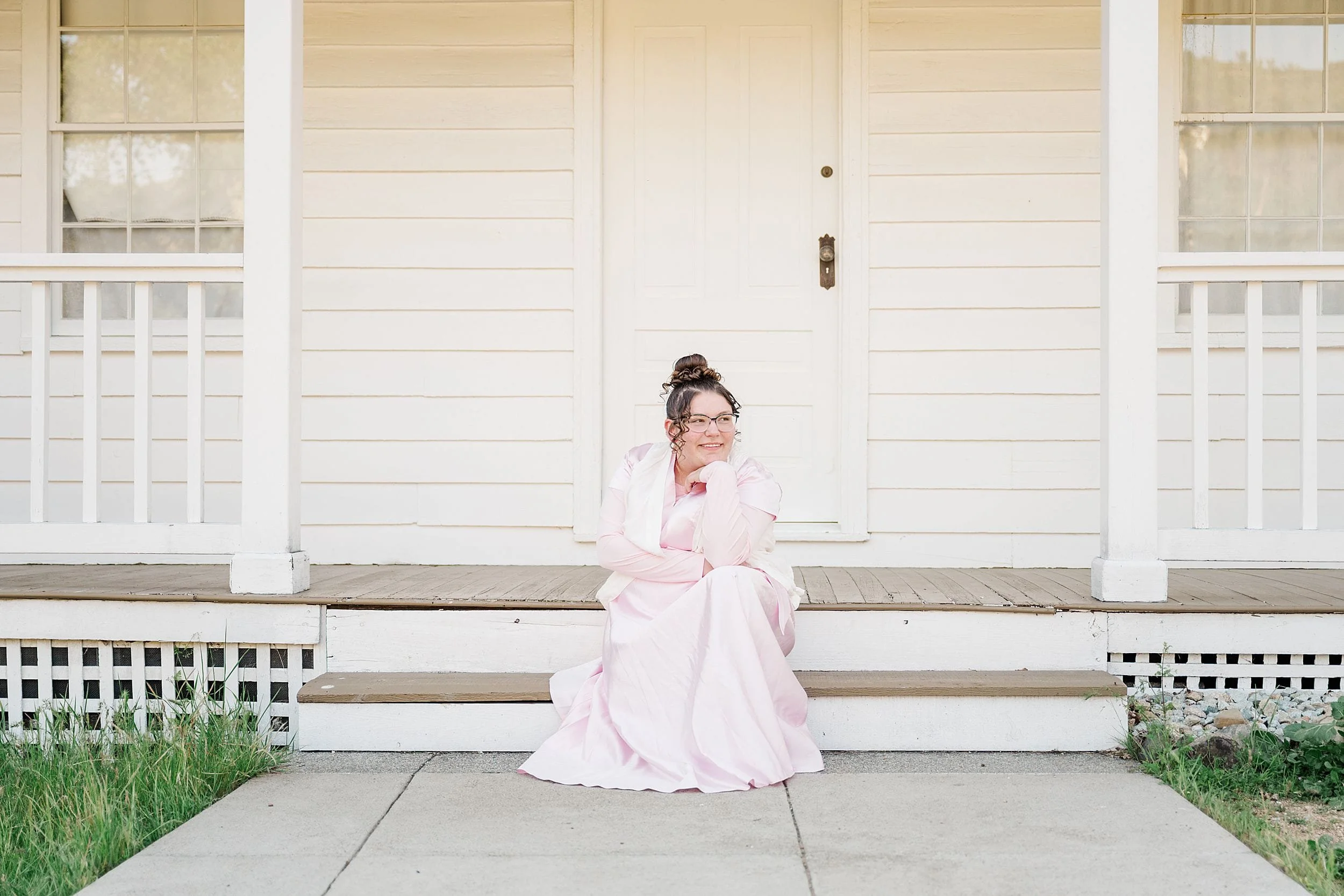 A woman in a pink dress sits smiling on the steps of a white wooden house with a closed door and windows. The mood is calm and content.