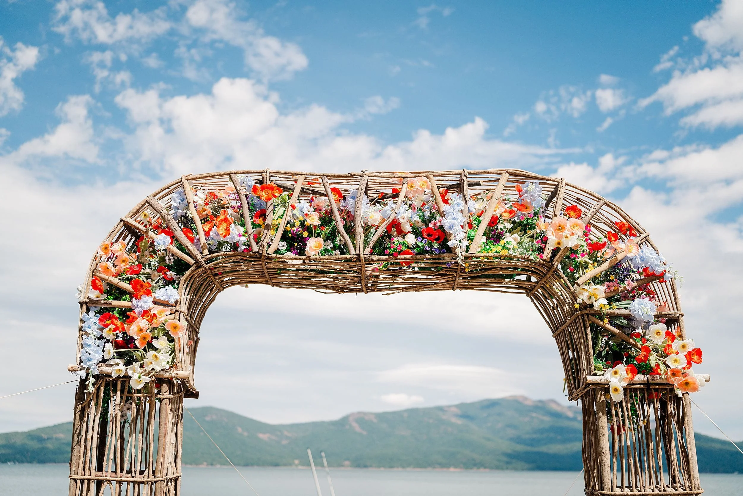 Rustic willow arch covered in colorful flowers stands beside a lake, framing green mountains beneath a bright blue sky; romantic and joyful.