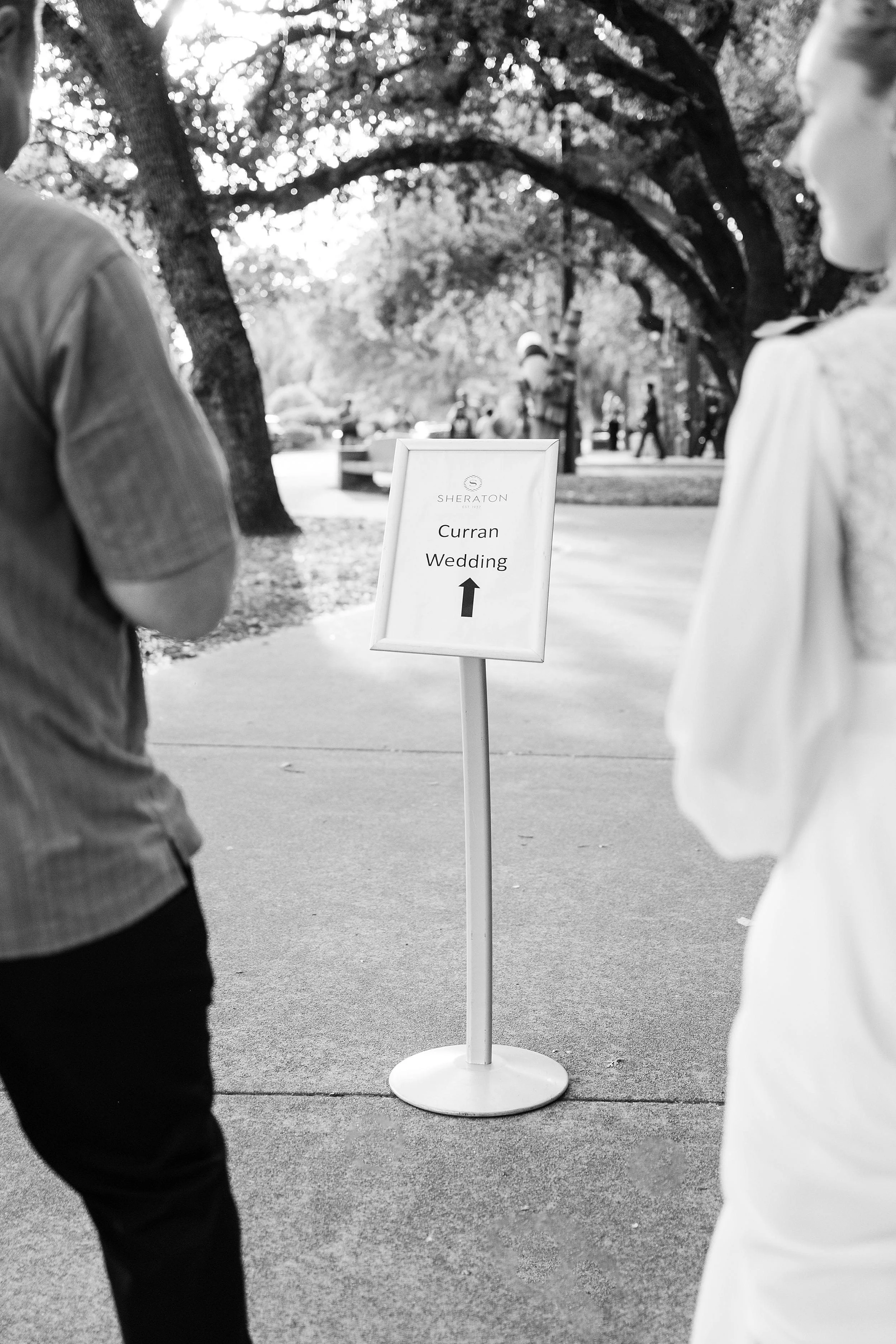 Black and white photo of a couple approaching a sign on a park path. The sign reads "Curran Wedding" with an arrow pointing forward. Trees line the path.