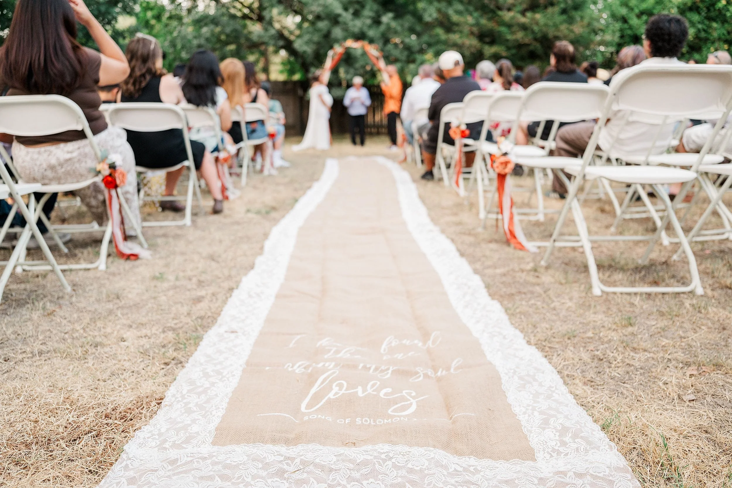 Outdoor wedding ceremony aisle with white chairs, floral arch, and lace-edged runner marked “love,” creating a romantic, joyful setting.