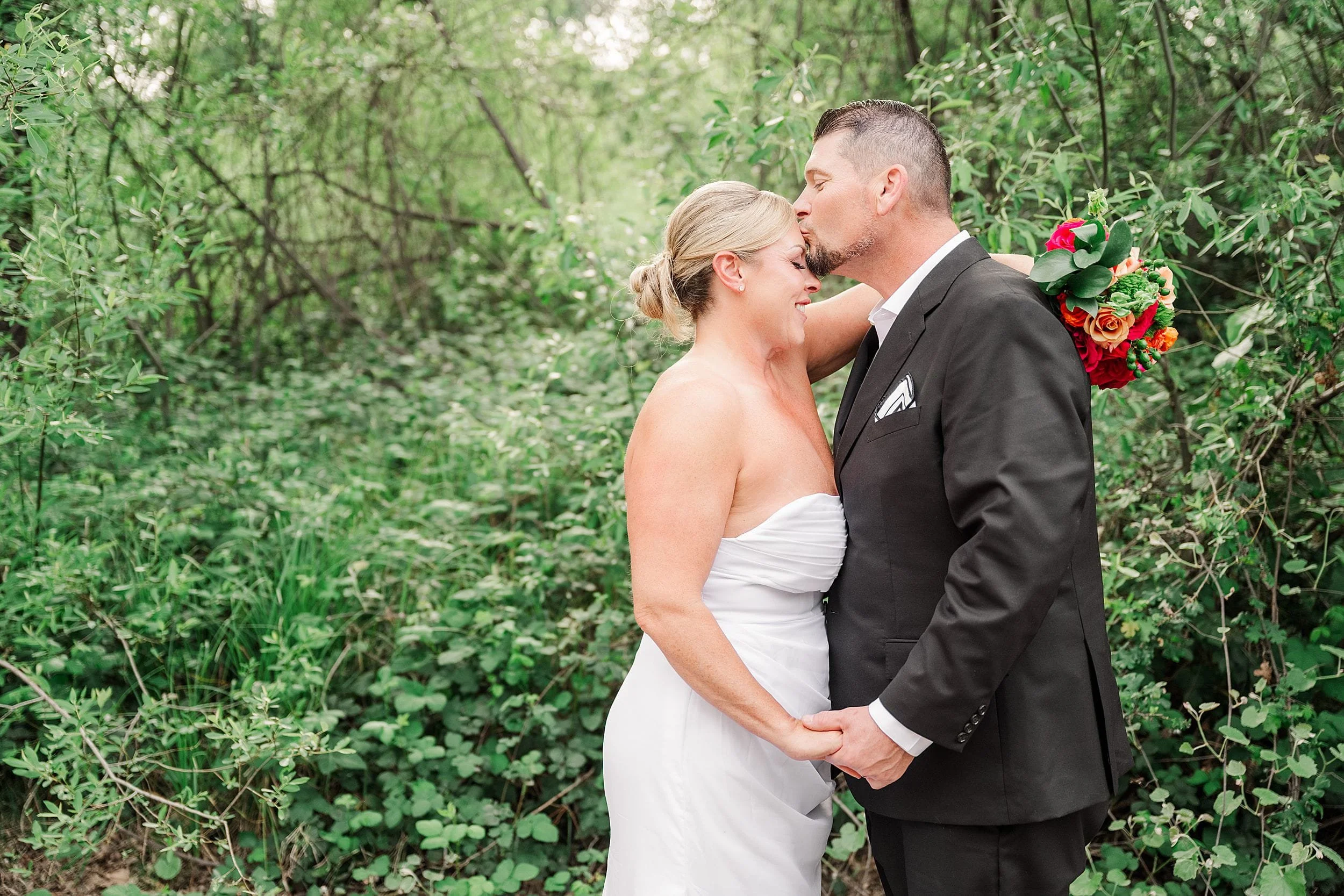 Bride in a strapless white dress and groom in a dark suit share a loving kiss on the forehead. They're surrounded by lush green foliage.