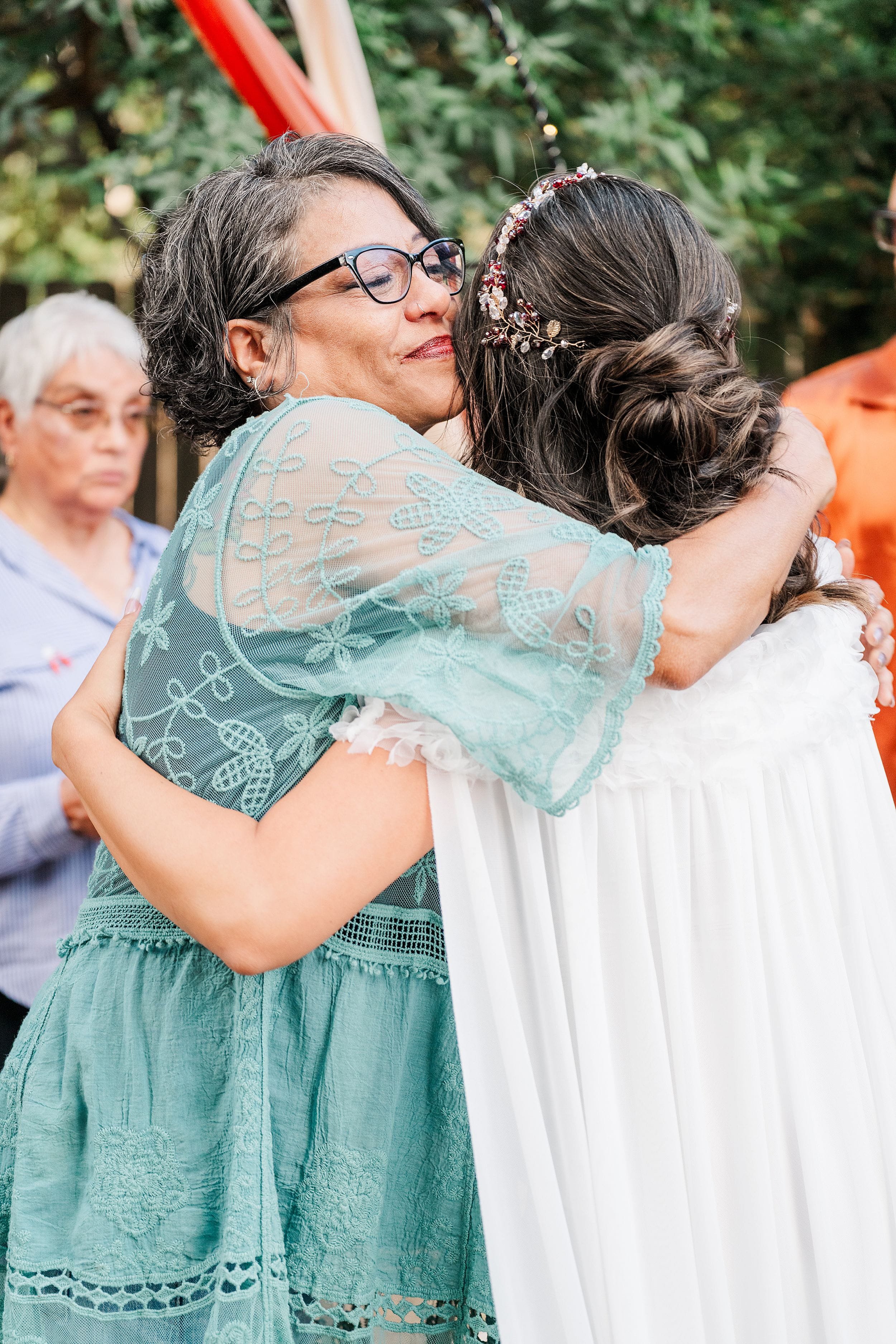An older woman in a teal dress warmly embraces a younger woman in a white dress, smiling at an outdoor celebration with guests nearby.