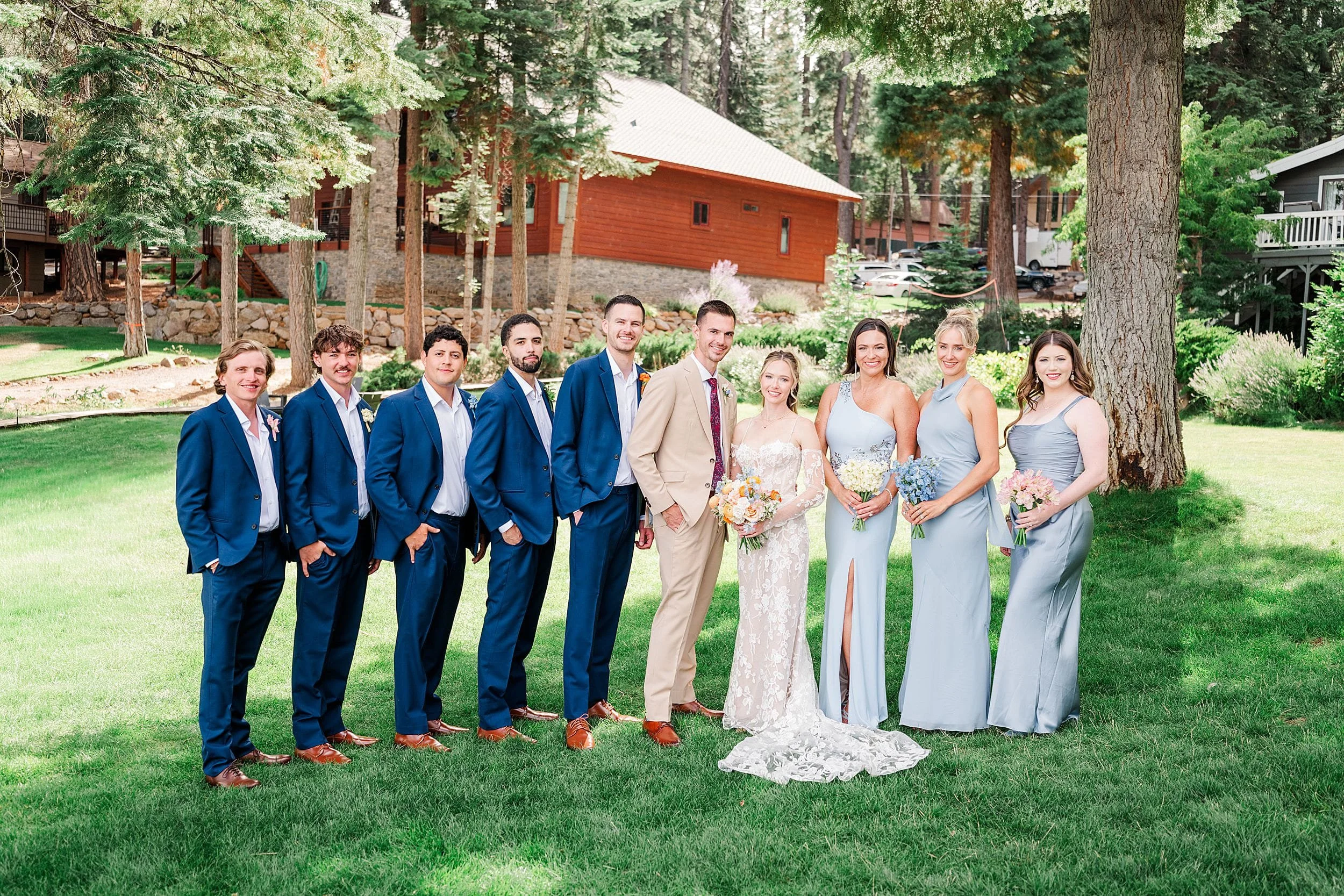 Wedding party poses on a sunny lawn at a forest lodge, with the groom and five groomsmen in suits and bride and three bridesmaids in pastel dresses.