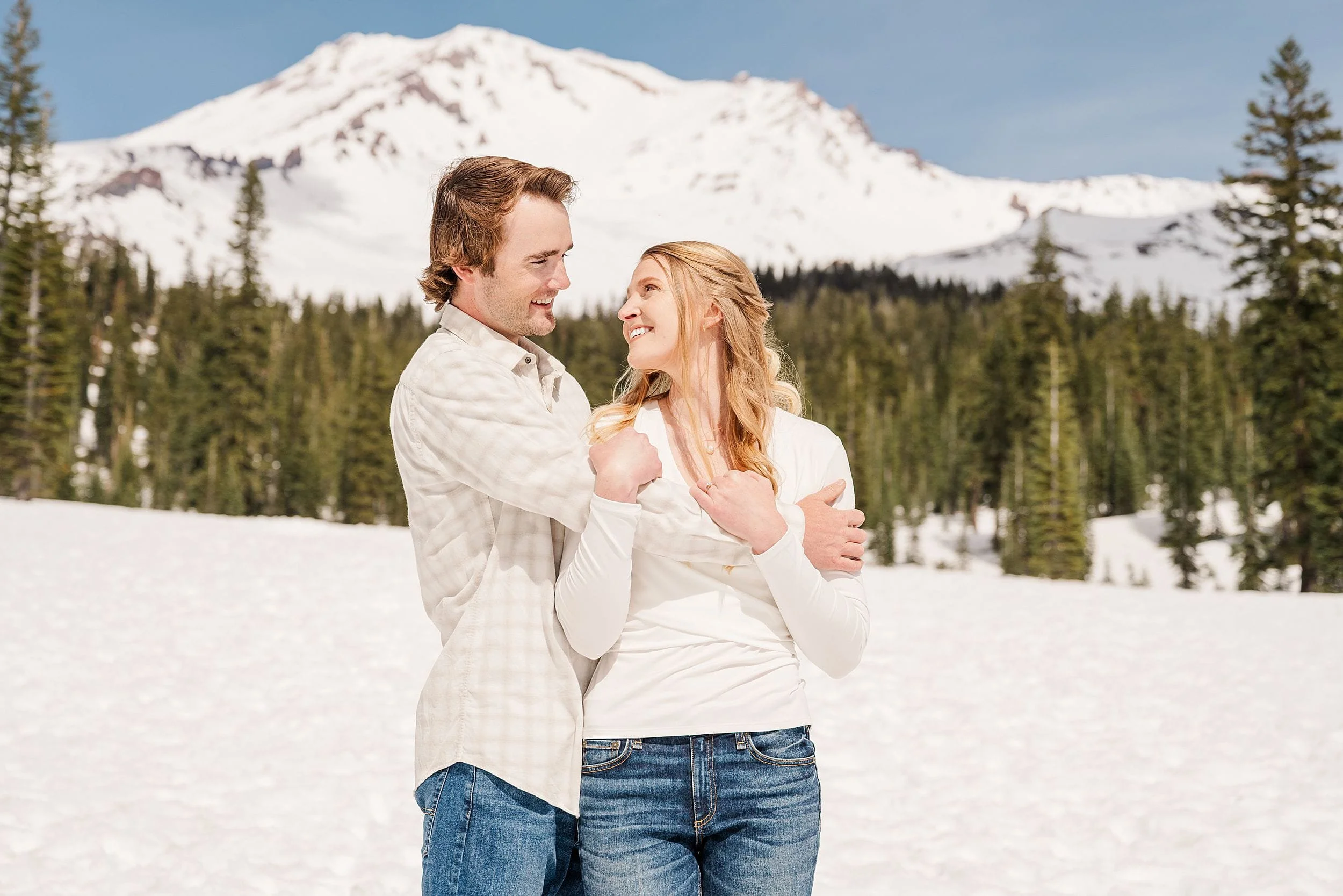 A couple stands in a snowy landscape, gazing at each other warmly. Behind them, a snow-capped mountain rises above evergreen trees under a clear blue sky.