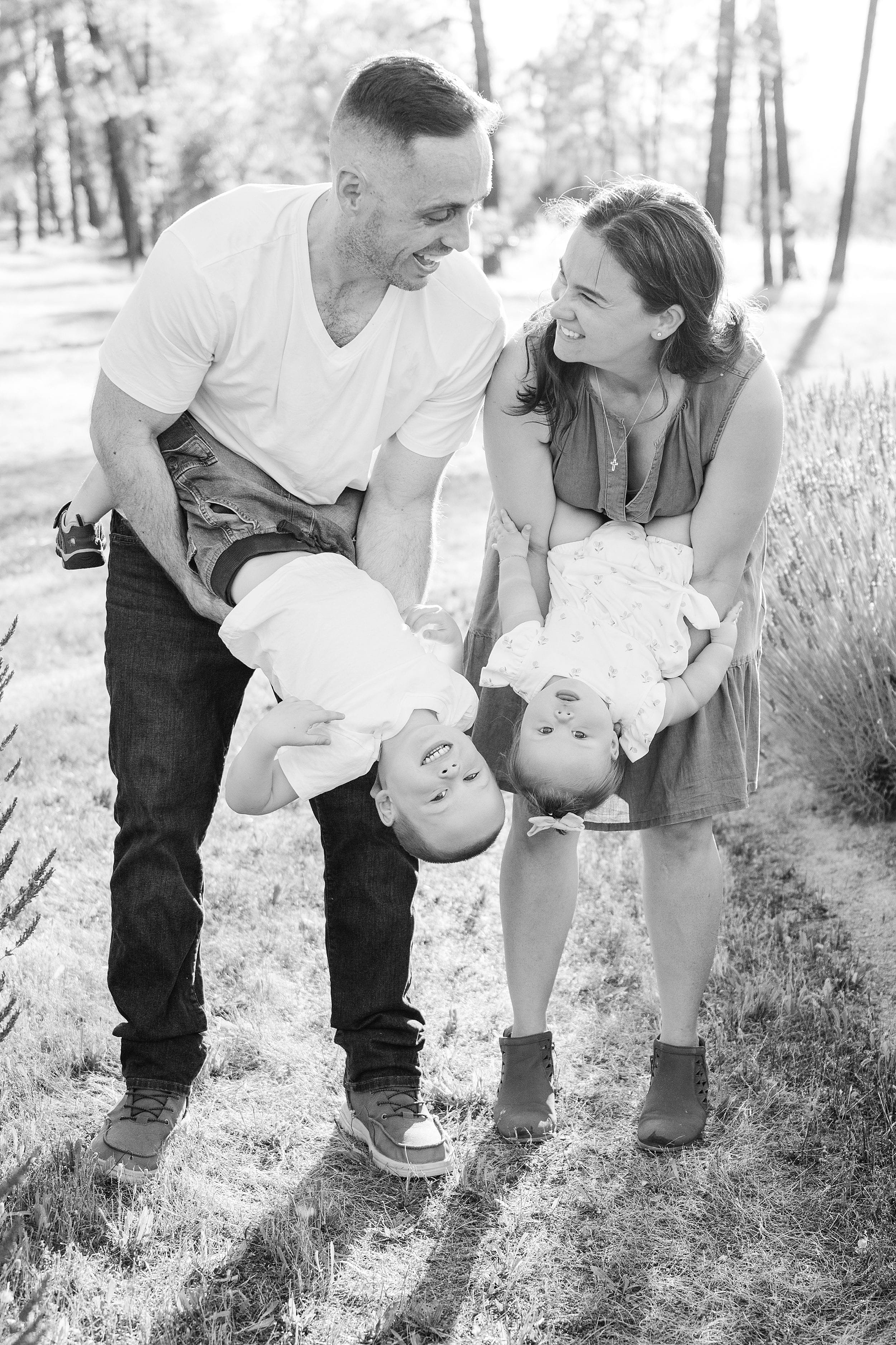 A joyful couple playfully hold their two young children upside down in a sunlit park, surrounded by tall trees. The scene is lighthearted and warm.