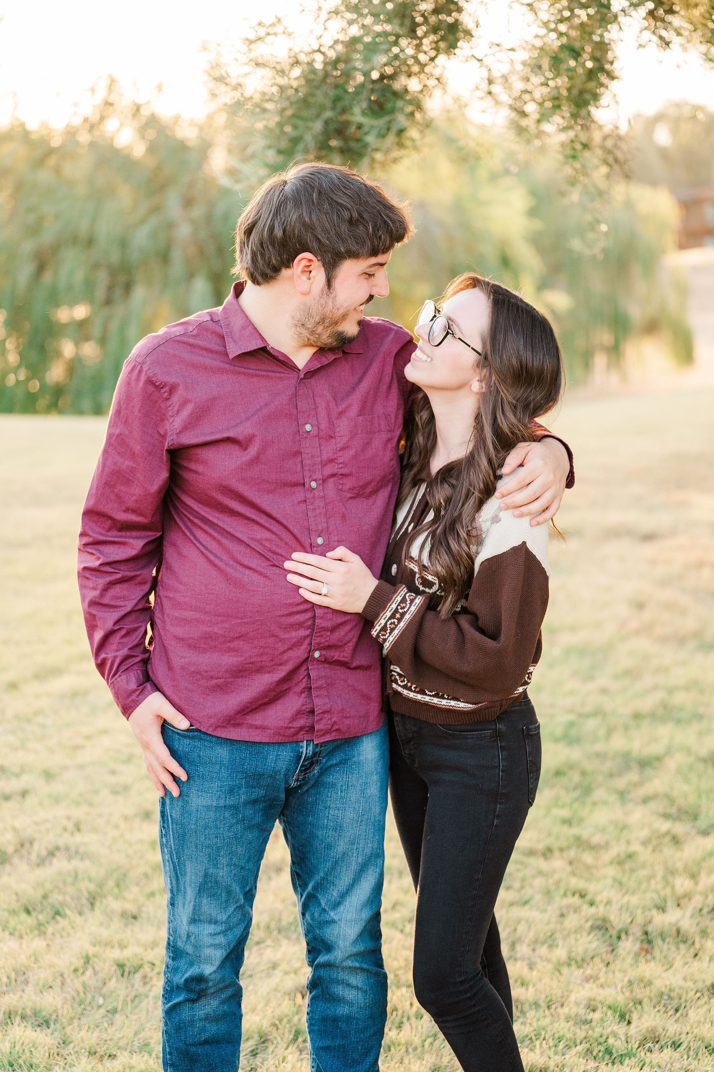 A smiling couple embraces in a sunlit field, gazing at each other amid tall grasses and trees; warm golden light creates a tender, romantic mood.
