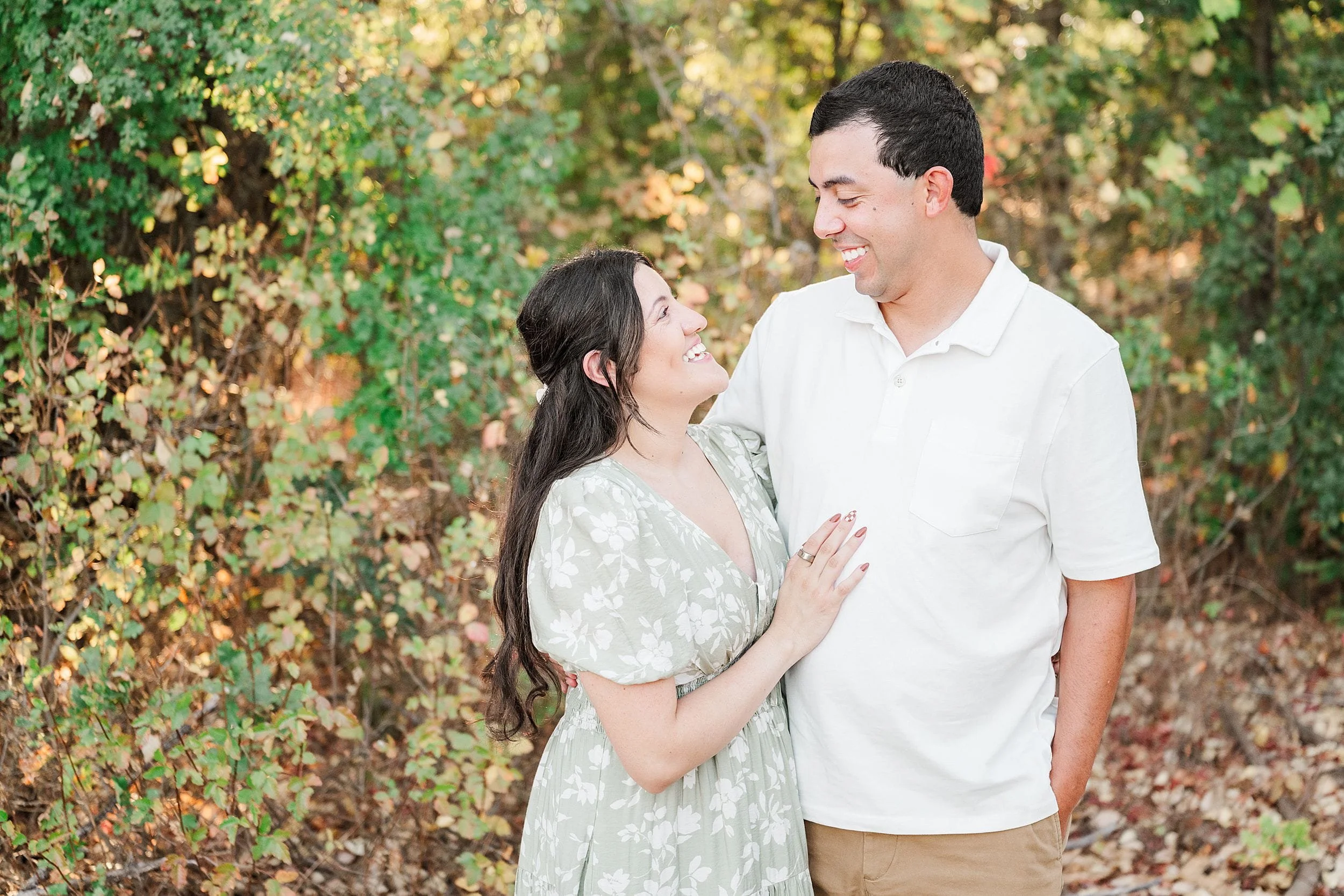 A smiling couple stands close together in a leafy autumn garden, gazing at each other as the woman rests a hand on his chest; warm, affectionate mood.