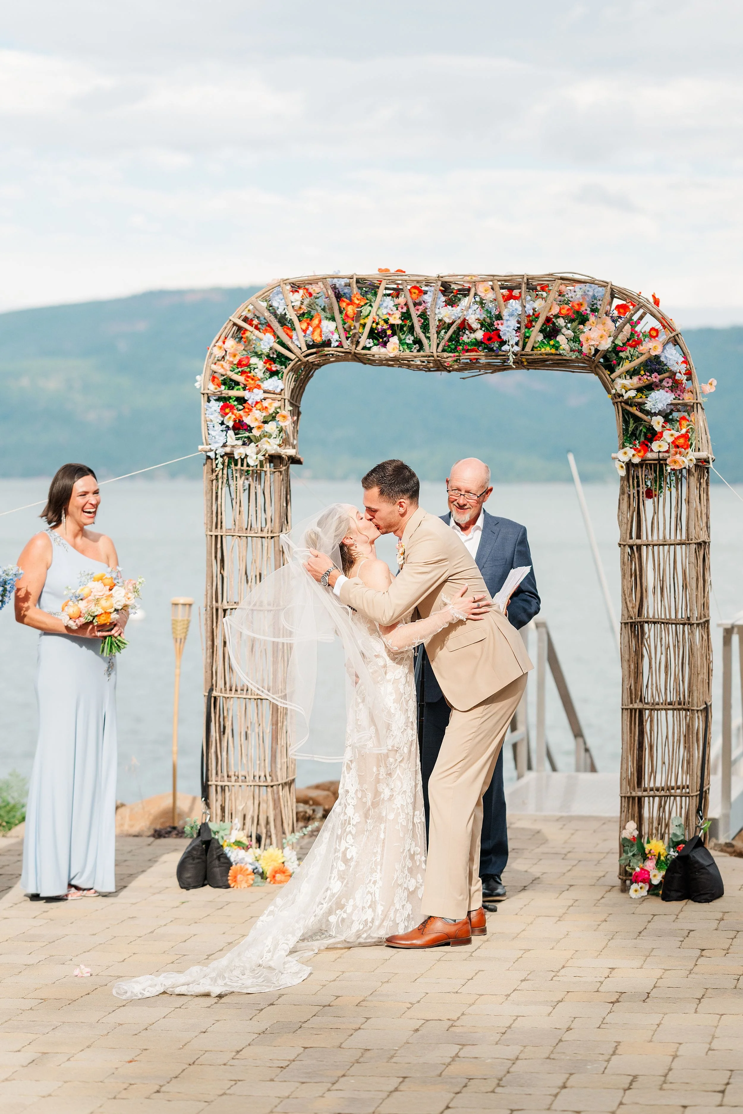 Bride and groom embrace beneath a flower-covered arch by a lake, while a bridesmaid and older man watch during an outdoor wedding ceremony.