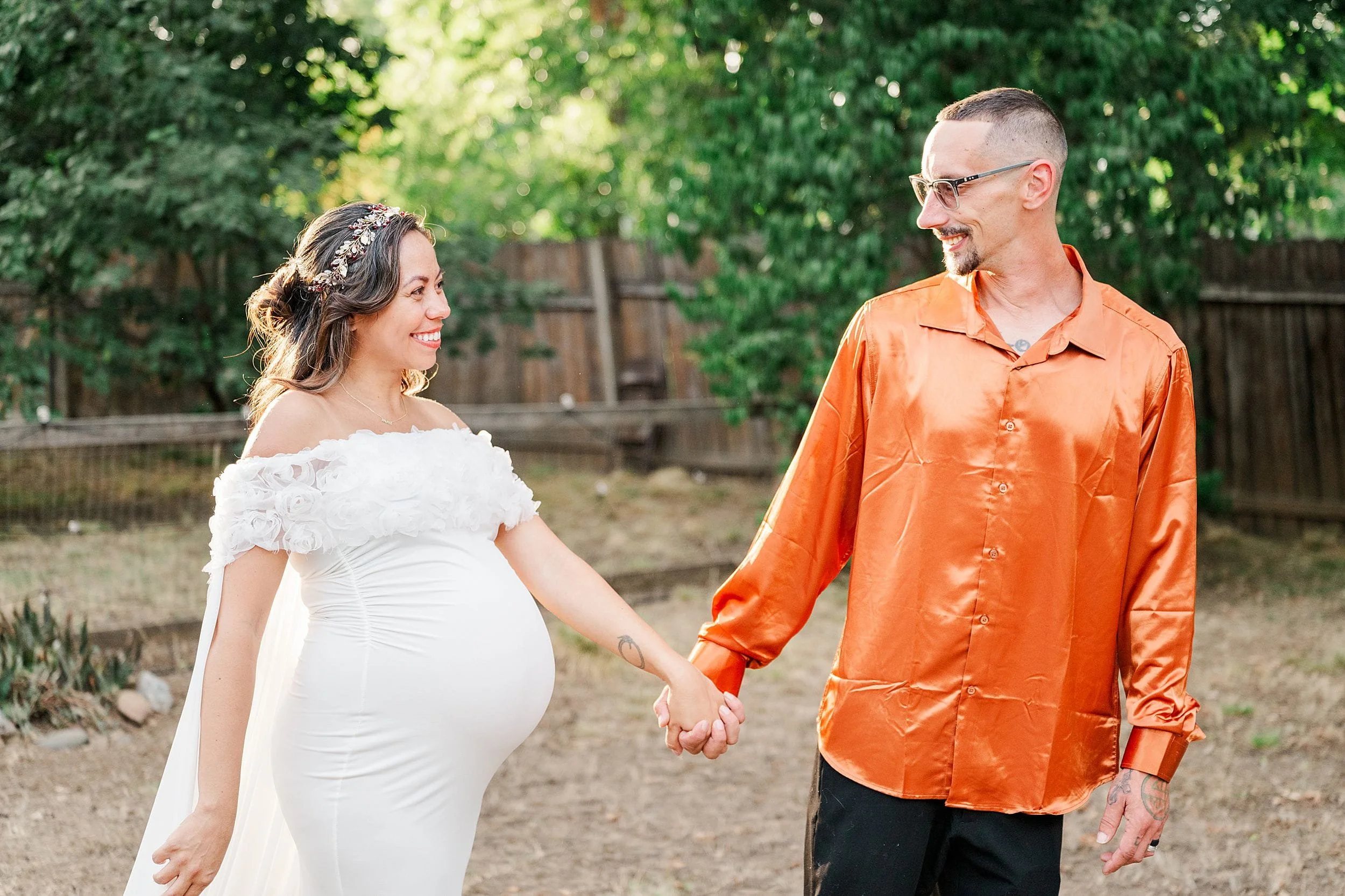 Pregnant woman in a white dress and smiling man in an orange shirt hold hands outdoors, sharing a joyful moment in a leafy backyard.