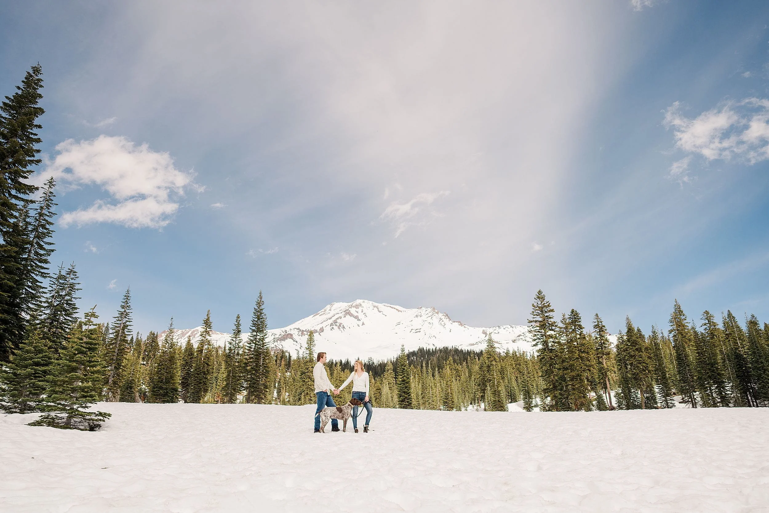 An engaged couple and their dog plays joyfully in a snowy landscape with tall evergreens and a snow-capped mountain in the background under a bright blue sky.