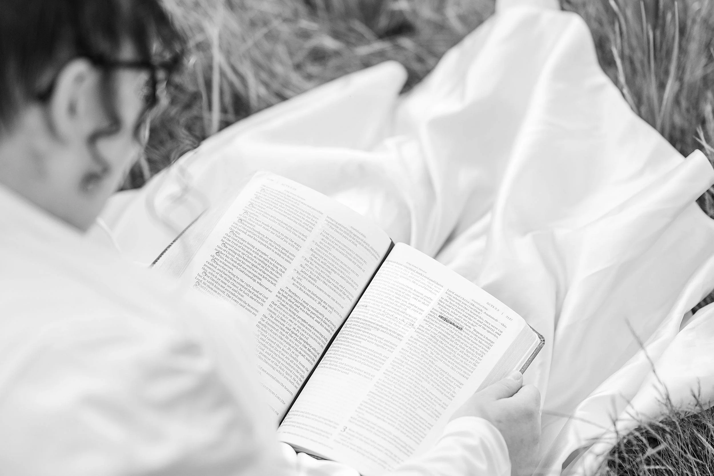 A person with glasses reads the bible outdoors, surrounded by grass. The image, in black and white, conveys a peaceful and contemplative mood.