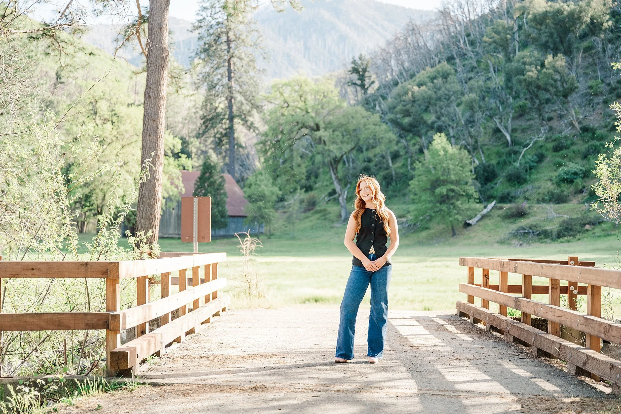 A person with long hair stands on a sunlit wooden bridge surrounded by lush green hills and tall trees, creating a serene and peaceful atmosphere.