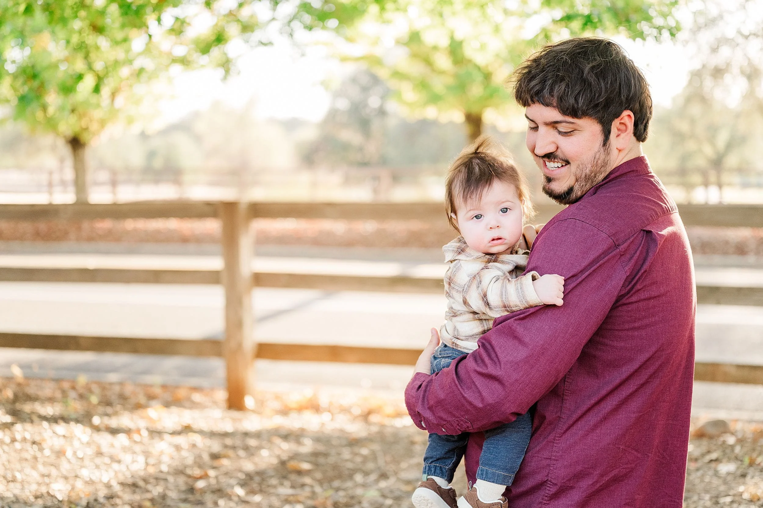 A smiling father in a burgundy shirt holds his baby outdoors beneath leafy trees, with a wooden fence behind them; a warm, tender family moment.