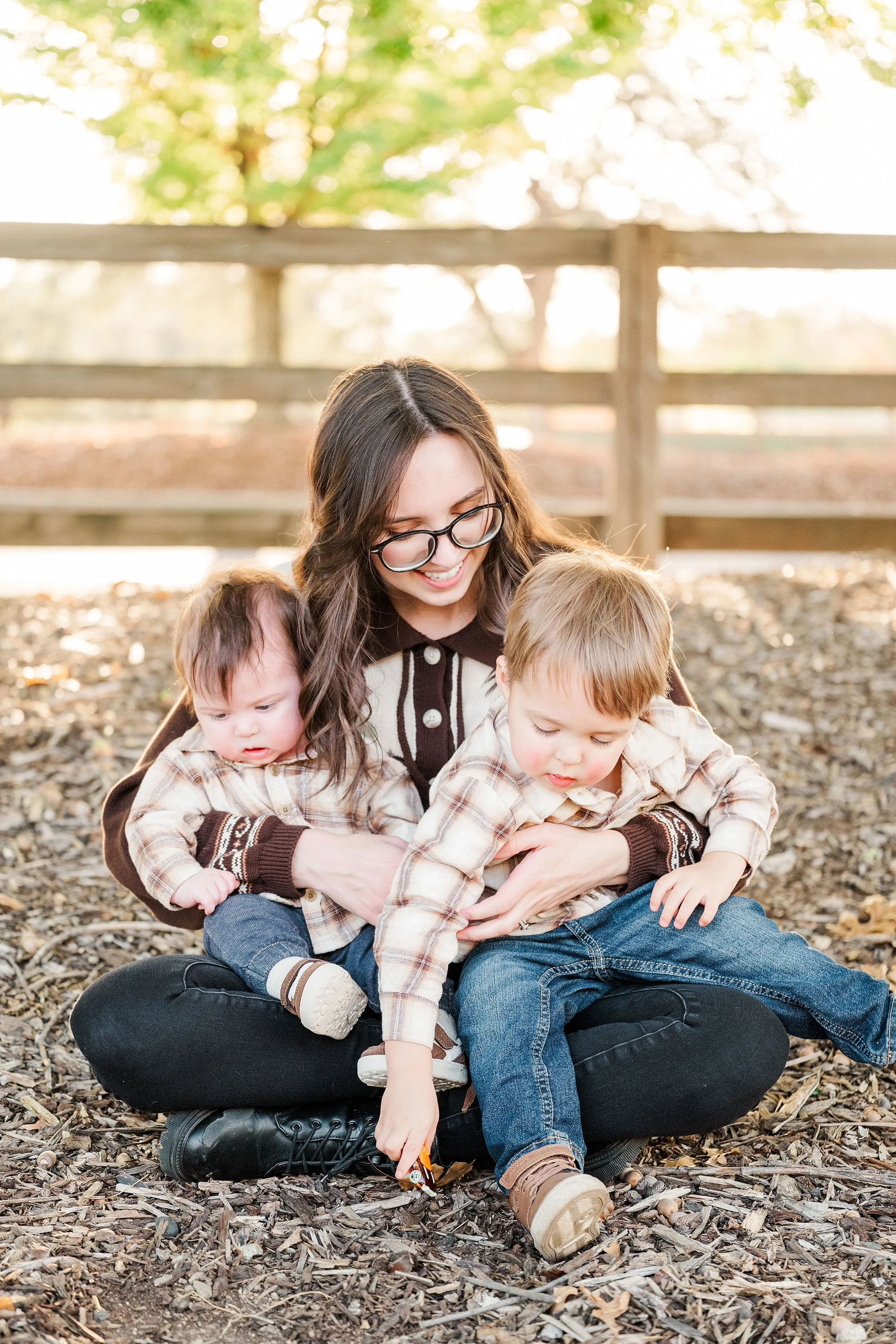 A smiling mother in glasses sits on leaf-strewn ground with two toddlers, helping one reach a leaf; a warm, sunlit family moment.