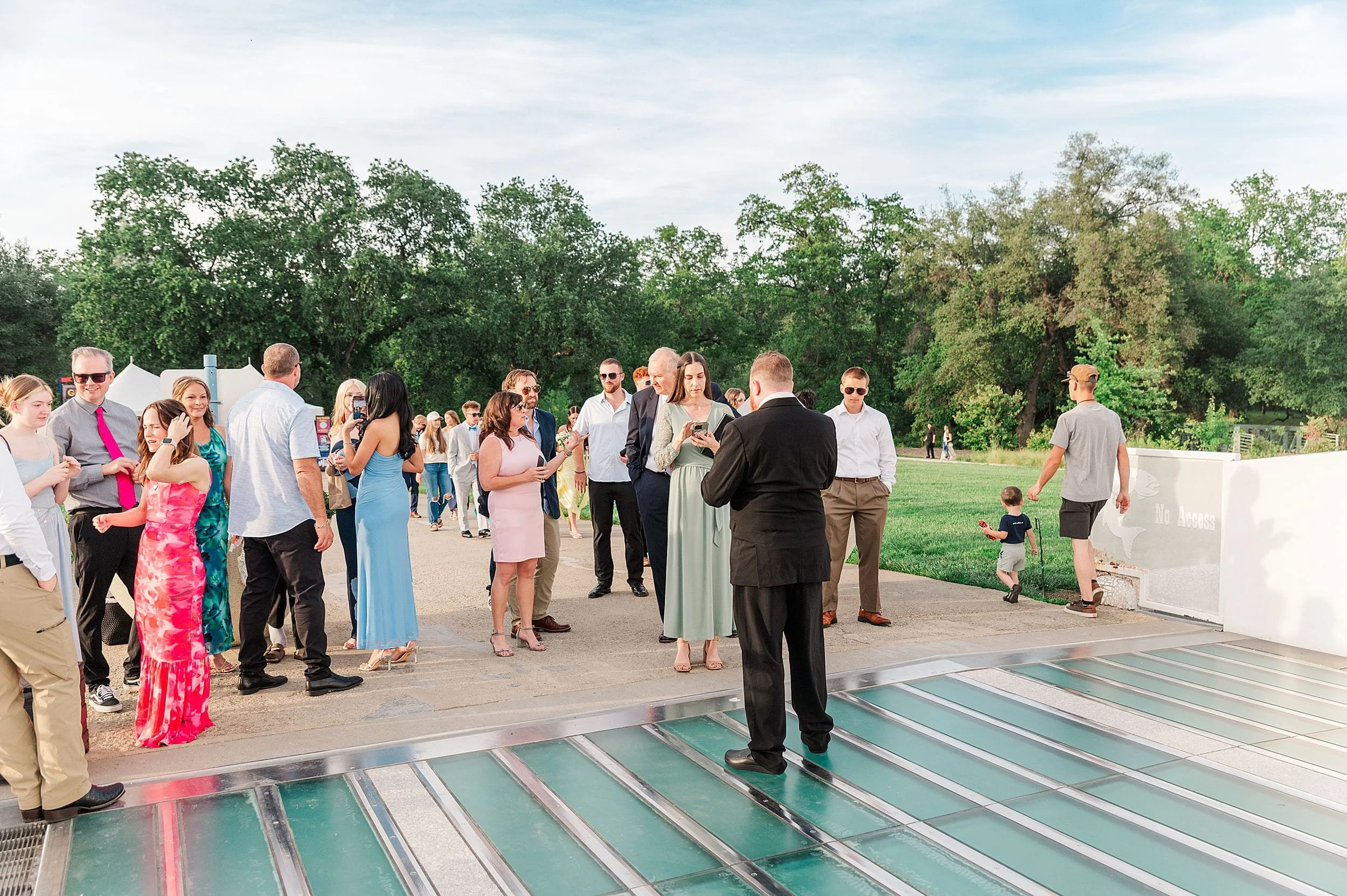 A group of people in formal attire gathers outdoors on a sunny day, engaging and mingling near a glass floor with lush trees in the background.