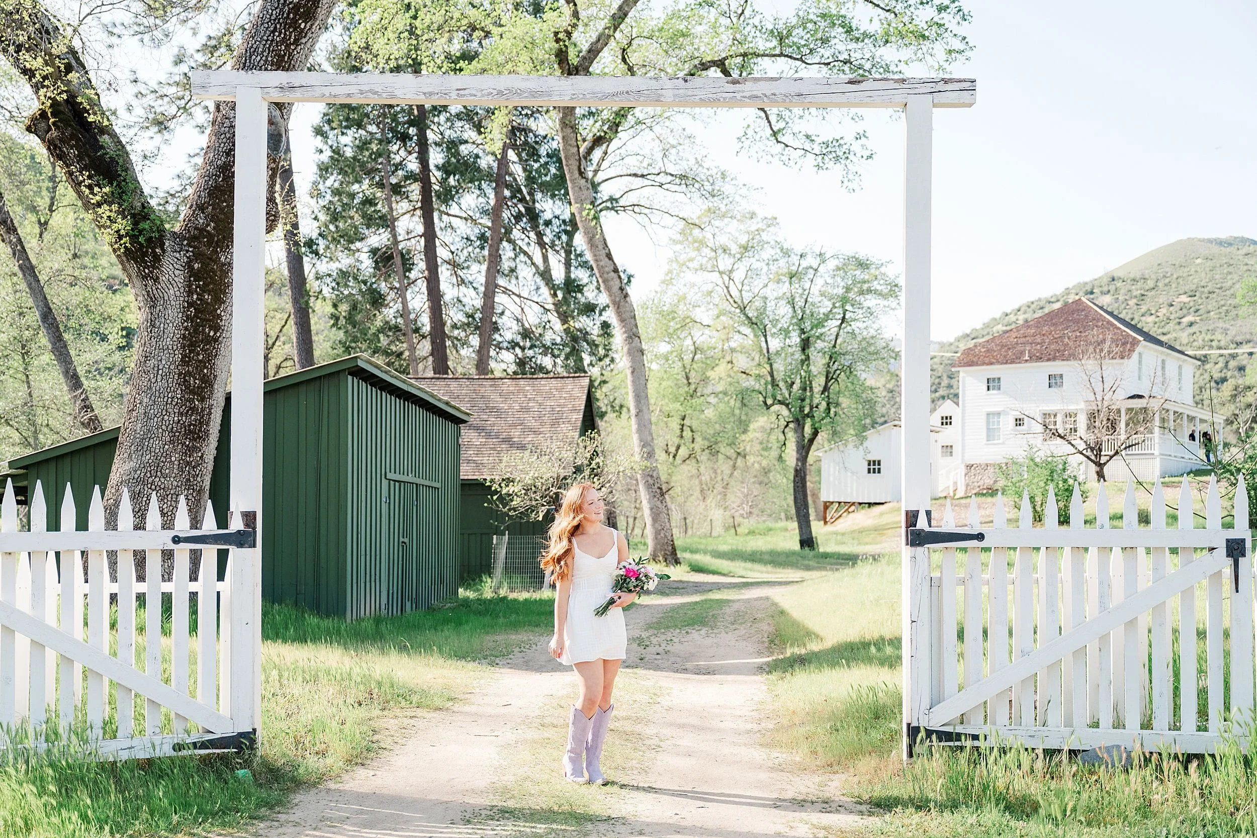 A woman in a white dress and lavender boots walks joyfully on a dirt path, holding flowers. She is framed by a white gate, with green trees and buildings behind her.