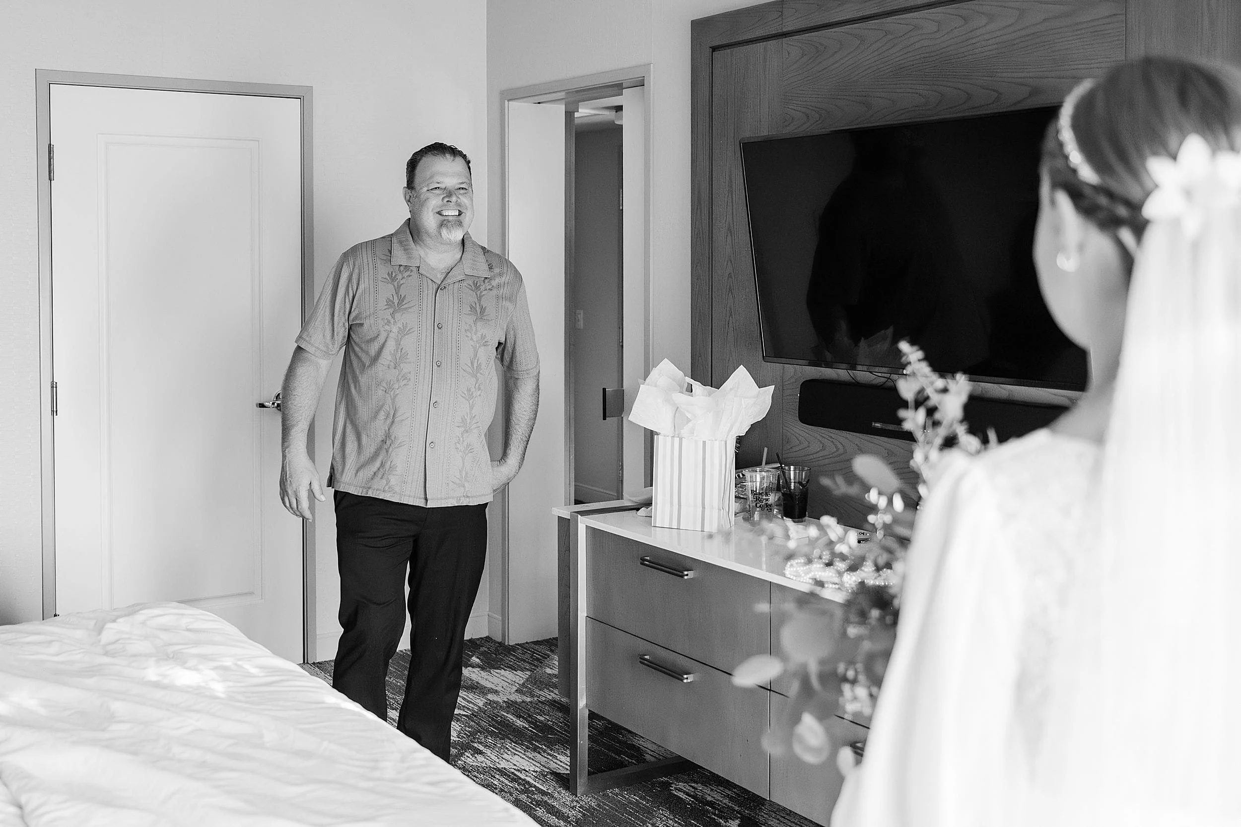 A smiling man enters a room, surprised, as a bride in a dress with flowers stands facing him. A wrapped gift sits on the dresser. Black and white photo.