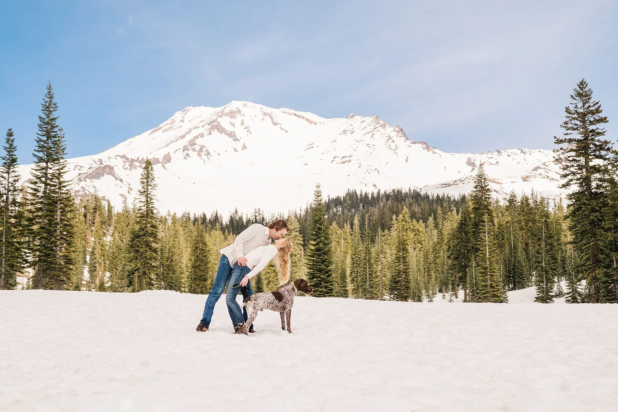A couple embraces on a snowy mountain slope with a dog at their feet. Evergreen trees surround them under a clear blue sky.